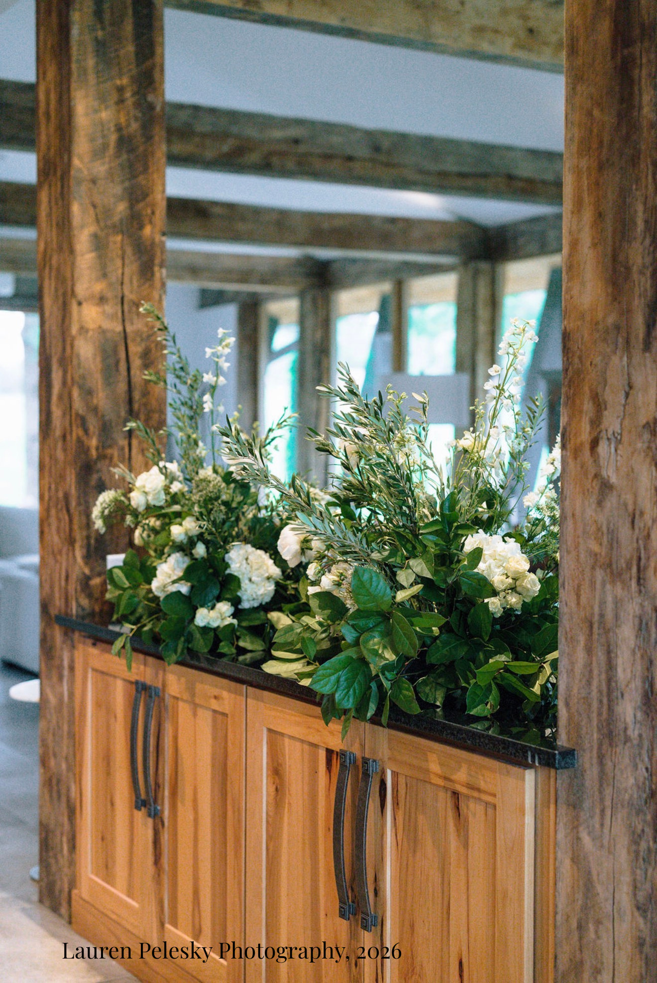 A wood cabinet is topped with beautiful flower clouds, filled with white roses, among a rustic room with wooden beams at a wedding dinner in Franklin, Tennessee.