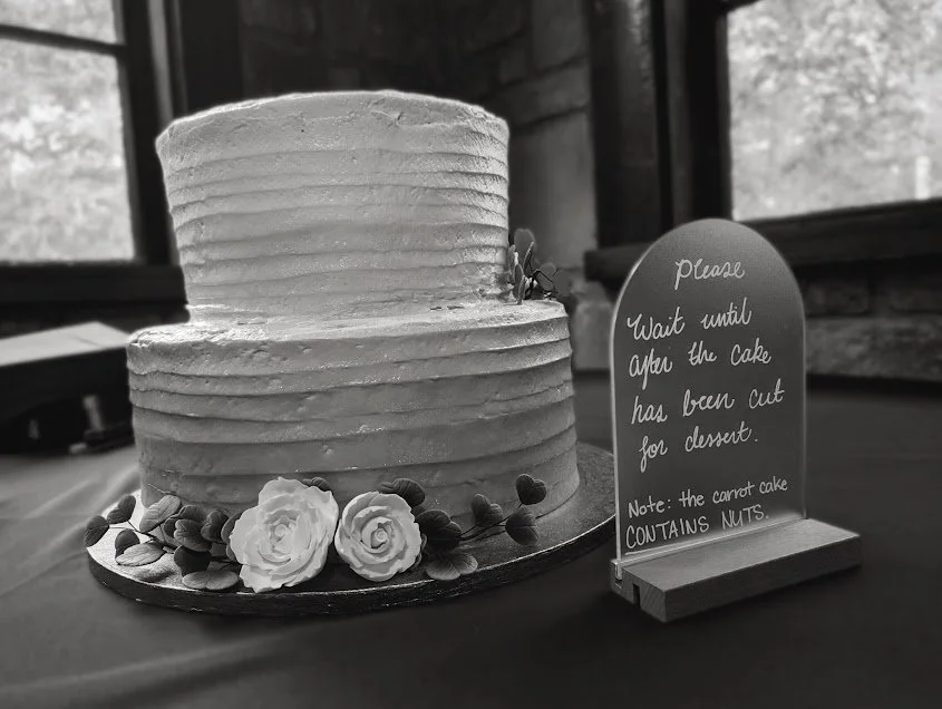 A two-tiered frosted cake decorated with flowers and leaves, placed on a table. A sign next to the cake requests to wait until the cake is cut for dessert and notes it contains nuts.