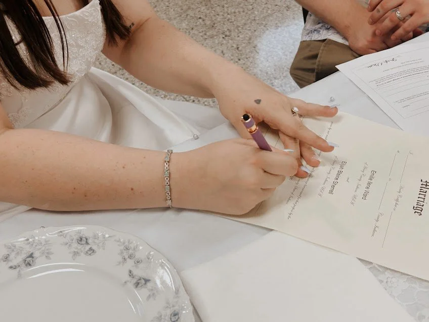 Person signing a marriage certificate at a wedding ceremony, wearing a white dress and bracelet, with wedding documents on a table.