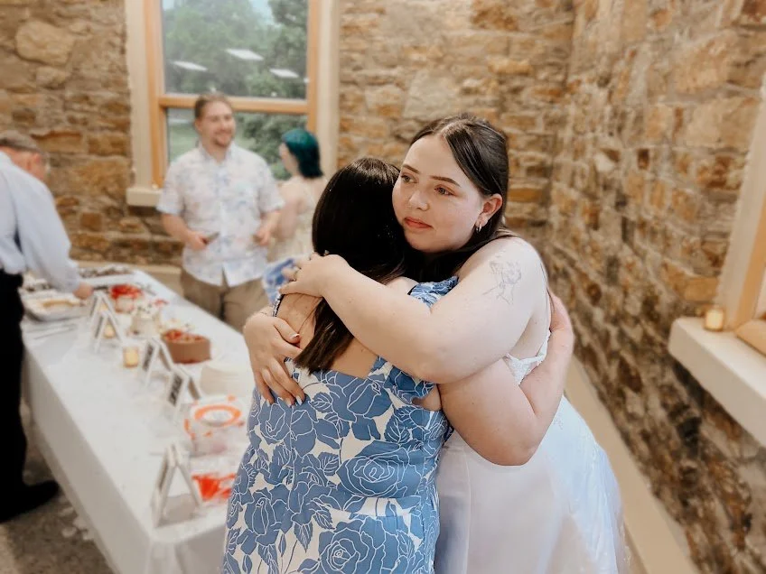 Two women hugging at a celebration, likely a wedding reception, with a table of food and people in the background.