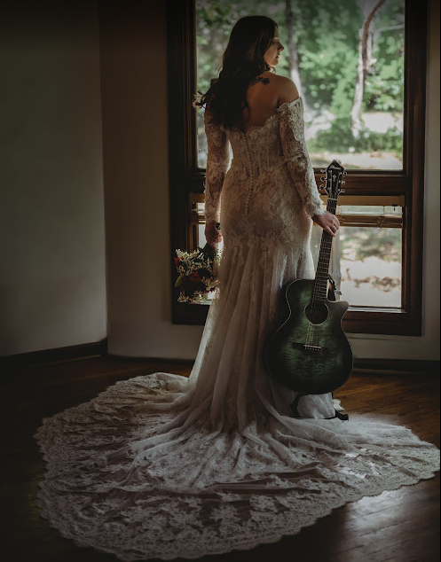 A woman in a wedding dress stands in front of a window, holding a blue acoustic guitar in one hand and a bridal bouquet in the other, at a wedding in Lebanon, Tennessee.