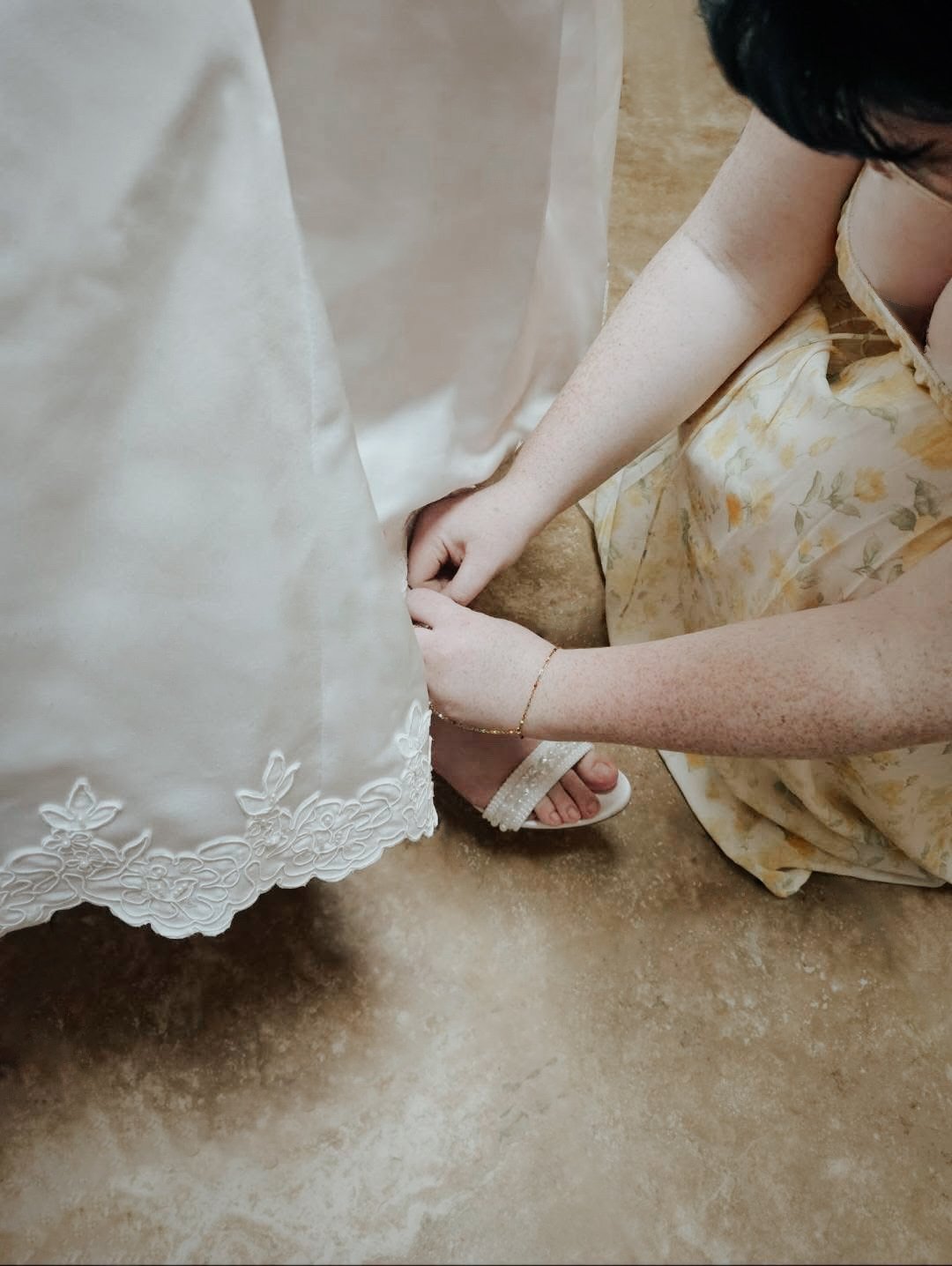 A bridesmaid in a yellow dress kneels down to adjust a bride's shoes.