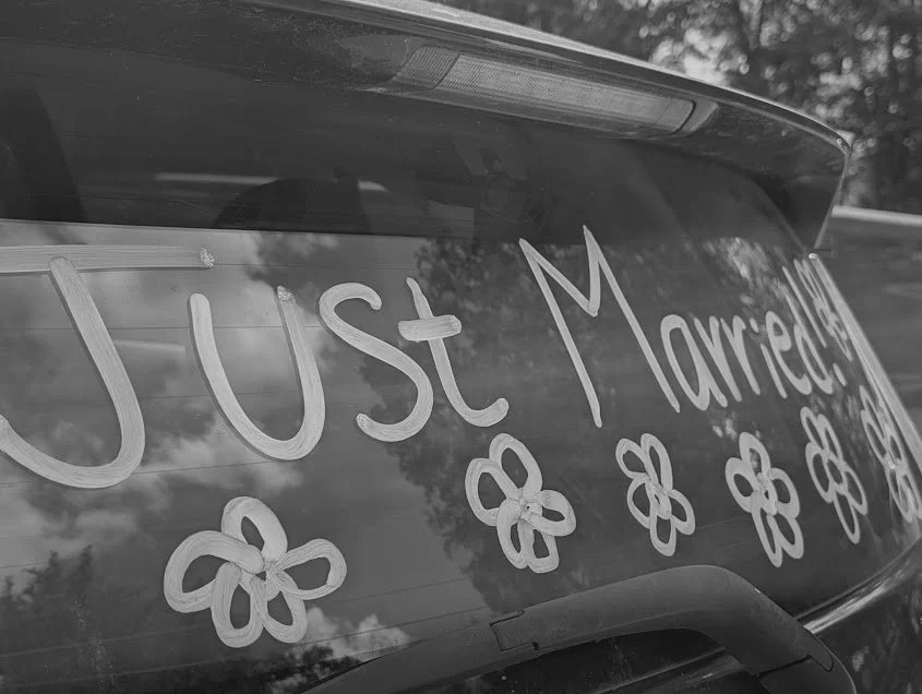 Black and white photo of a car's rear window decorated with handwritten message 'Just Married' and surrounded by flower illustrations.