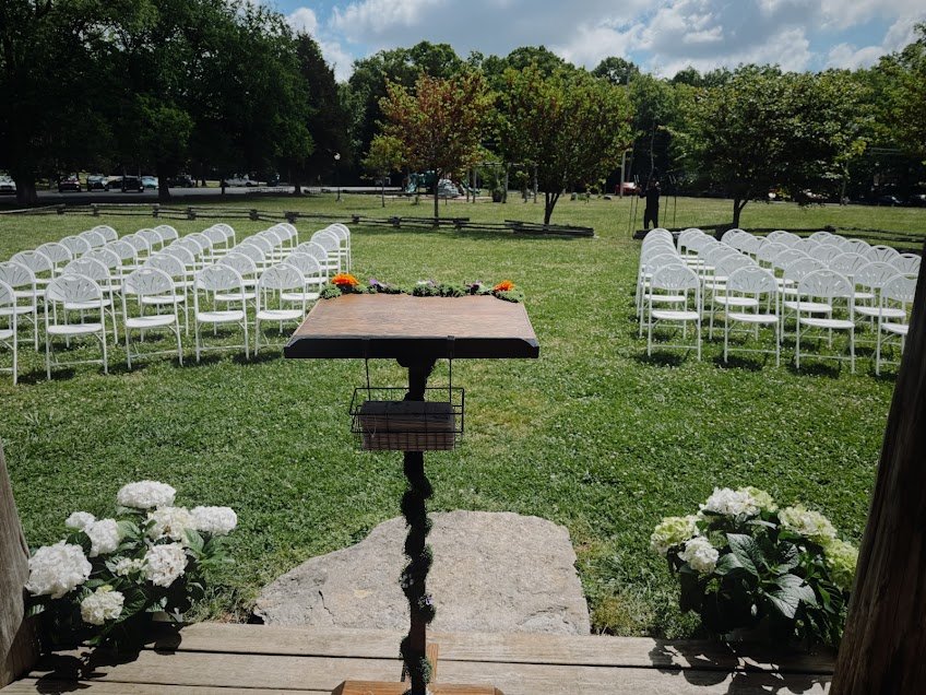 Outdoor wedding ceremony setup with white chairs facing a small table decorated with flowers, greenery, and trees in the background.