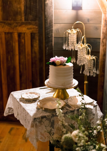 A small wedding cake sits atop a cake table with a lace table cloth. Beside the table is a chandelier style lamp and a flower cloud.