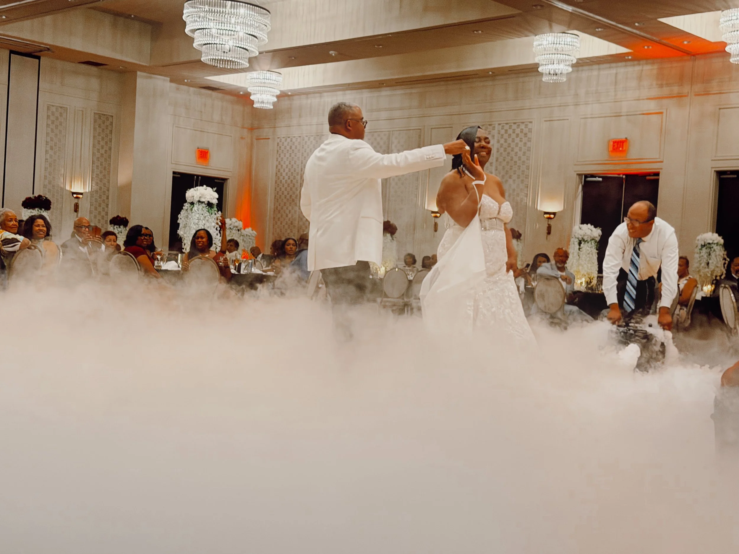 A bride and groom share their first dance in a beautifully decorated ballroom, as a fog machine fills the dance floor with fog. The groom wears a white suit jacket while the bride wears a lace, sleeveless ballgown with a sweetheart neckline.