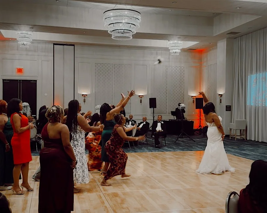 A bouquet toss takes place in an elegant hotel ballroom with hanging crystal chandeliers. The bride's bouquet flies through the air as bridesmaids and guests lunge and jump to catch the bouquet as the bride turns around to watch.