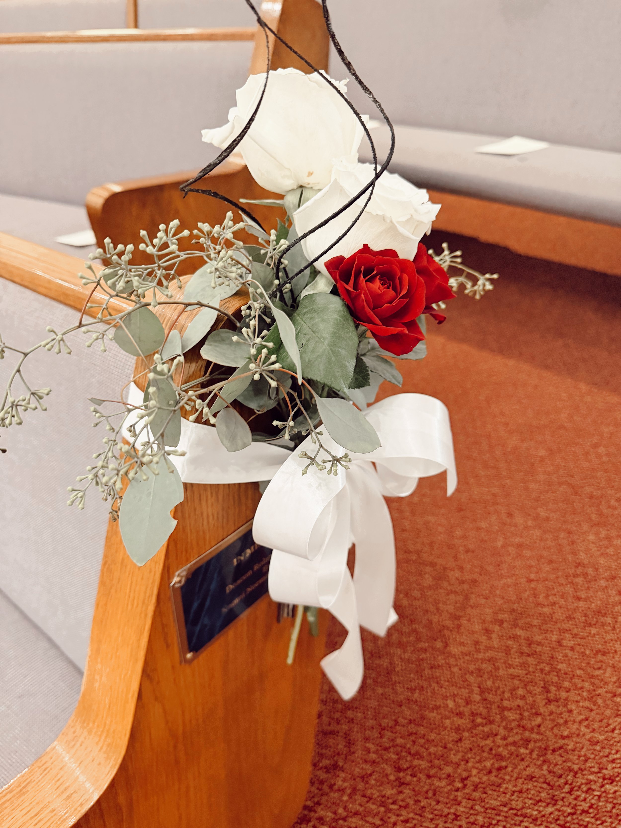 A floral aisle marker containing red and white roses and sage greenery is tied around a church pew with a white ribbon at a wedding ceremony at a Baptist church in Nashville, Tennessee.