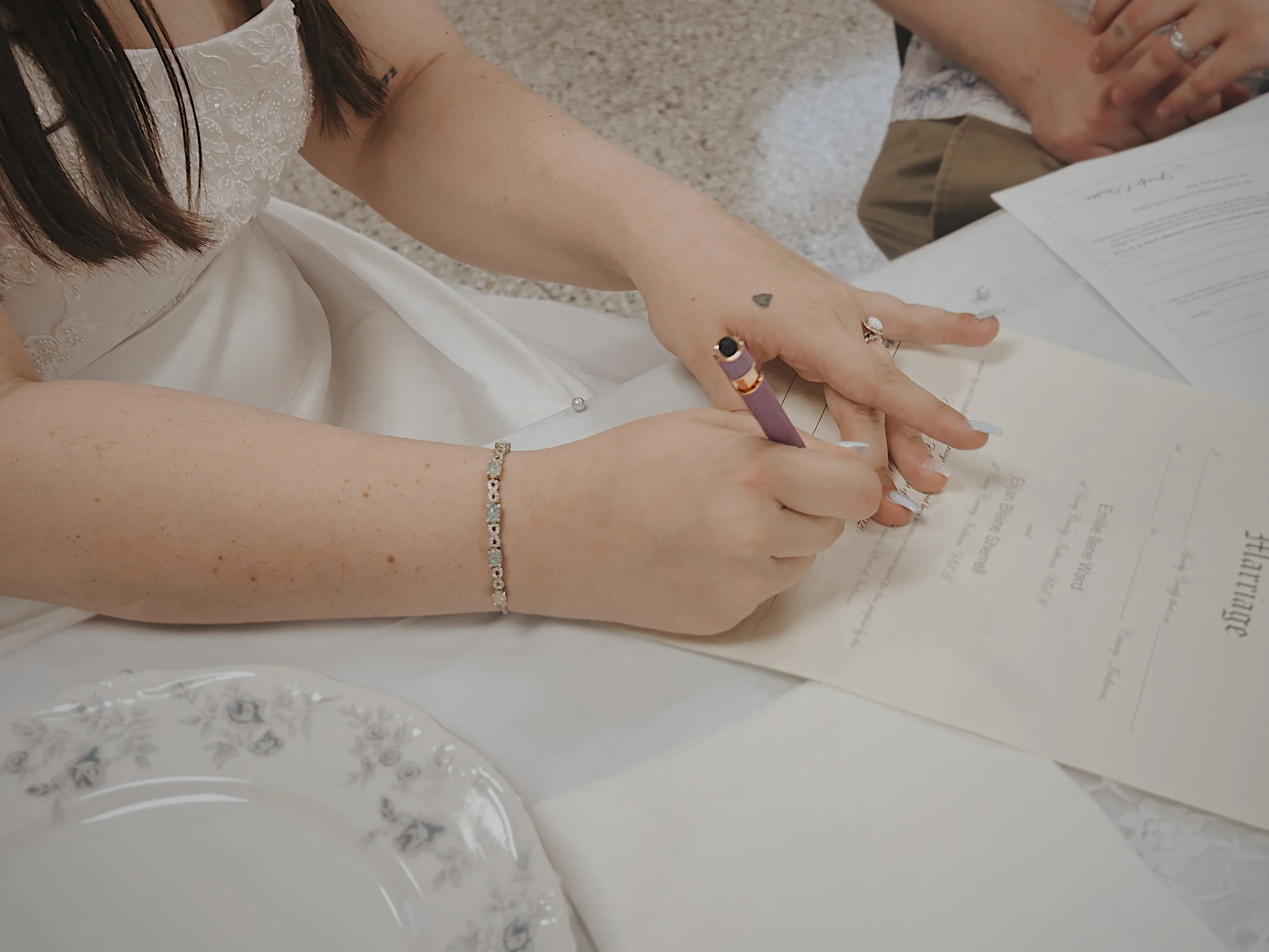 A woman in a white wedding dress, wearing an opal engagement ring, signs her marriage license at a wedding in New Castle, Indiana.