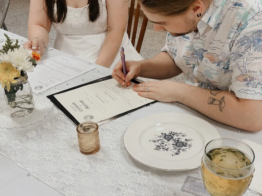 A man signing a marriage certificate at a table decorated with flowers, a lace tablecloth, and glassware, with a woman standing nearby.