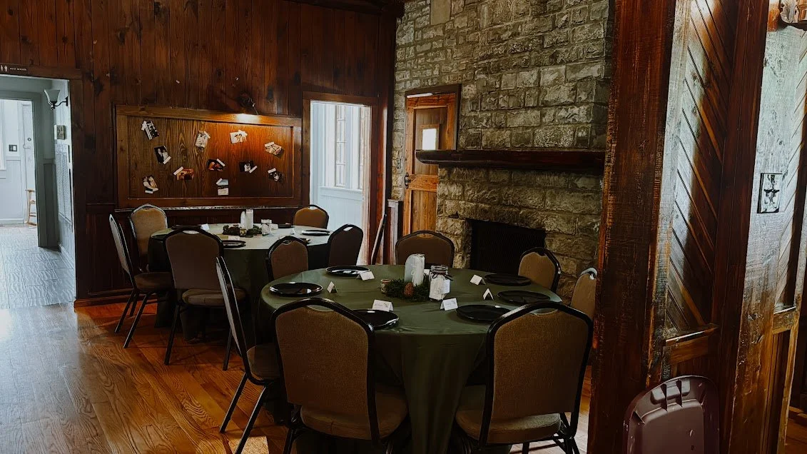Dining room with round table set for a meal, surrounded by chairs, stone fireplace, and wood-paneled walls.