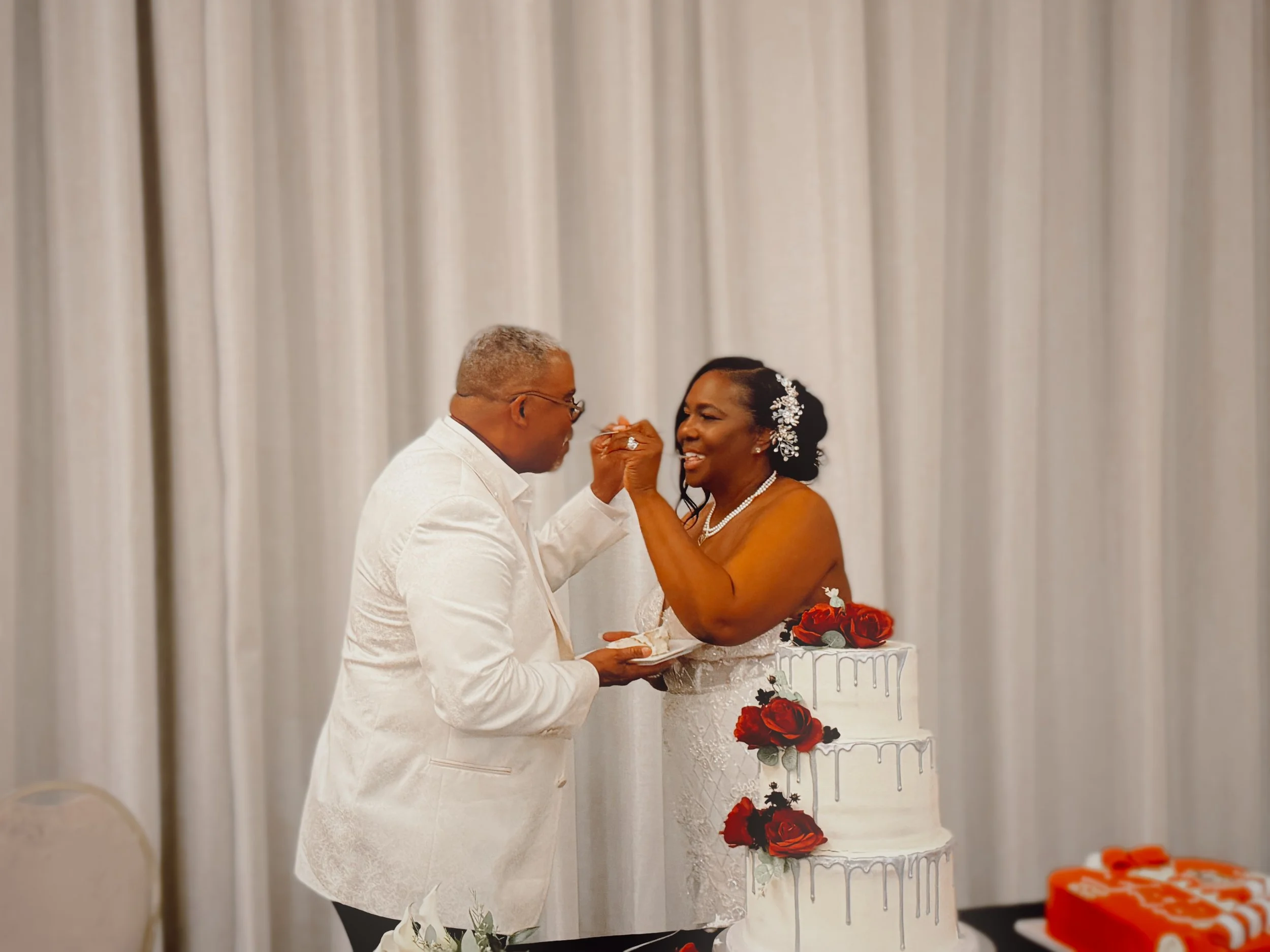 A bride and groom stand behind a three tier wedding cake decorated with red roses and purple flowers. They are smiling while feeding each other the first slice of cake.