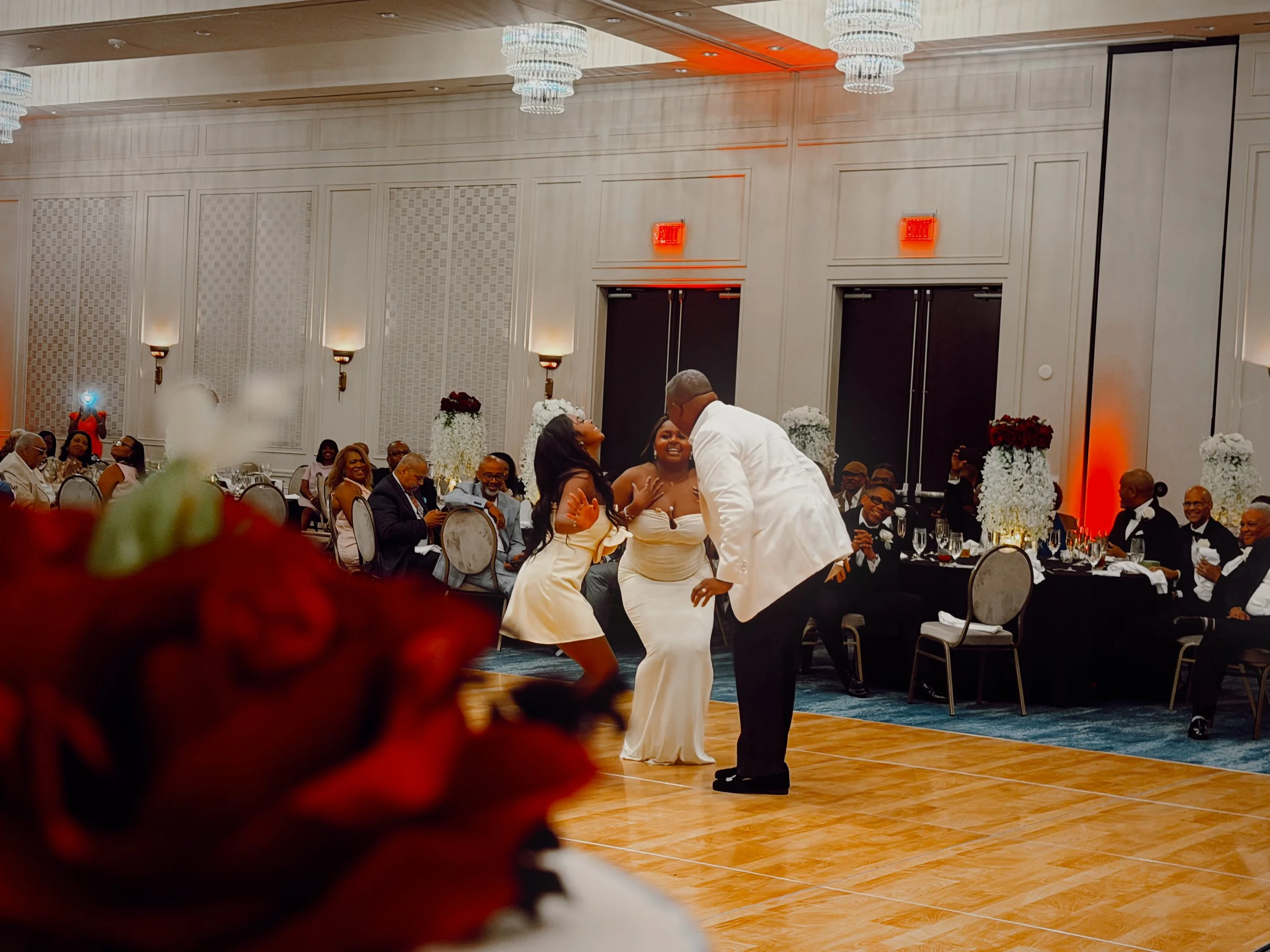 The groom dances with his daughters, both wearing butter yellow dresses, at a wedding reception in a ballroom decorated with white and red rose centerpieces, black tablecloths, and candles in Nashville, Tennessee.