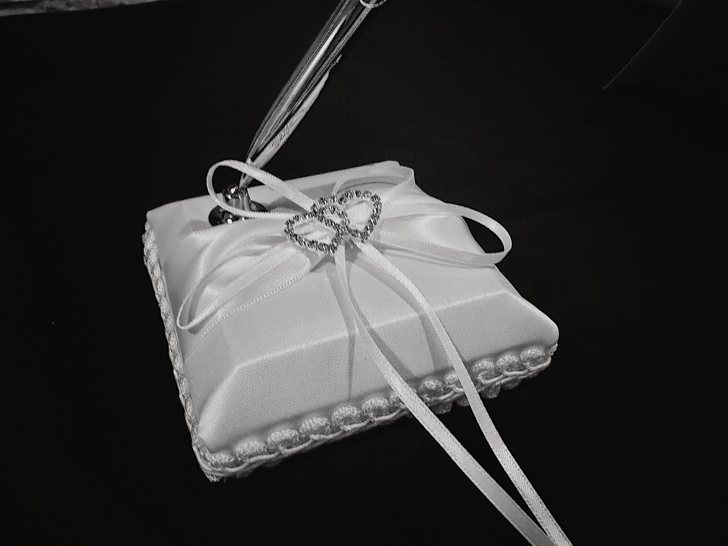A white satin cushion holds a fountain pen for guest book signing. The satin cushion has a white ribbon tied in a bow and two jewel encrusted hearts.