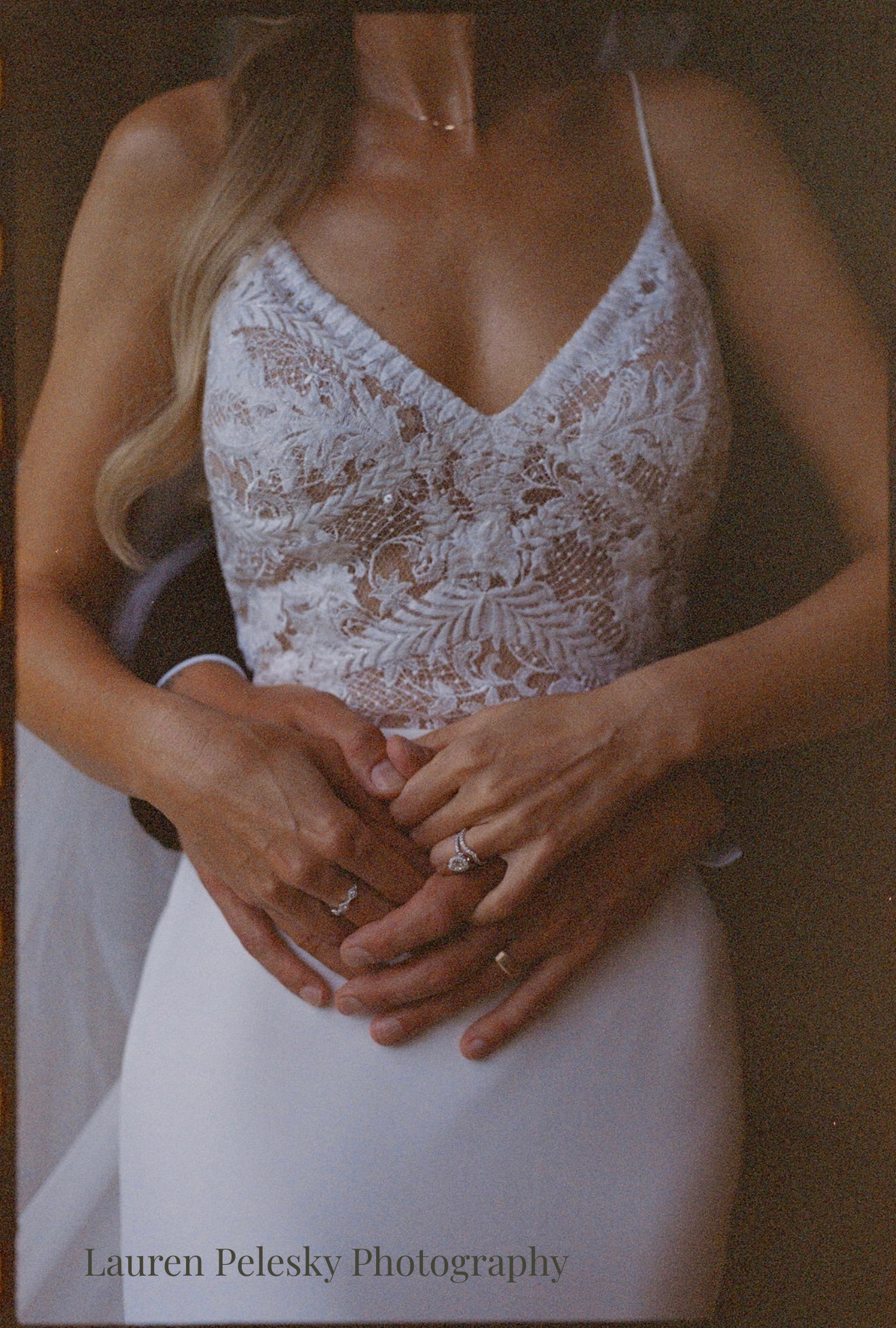 A groom hugs his bride from behind, interlocking hands over her lace gown, displaying their wedding rings.