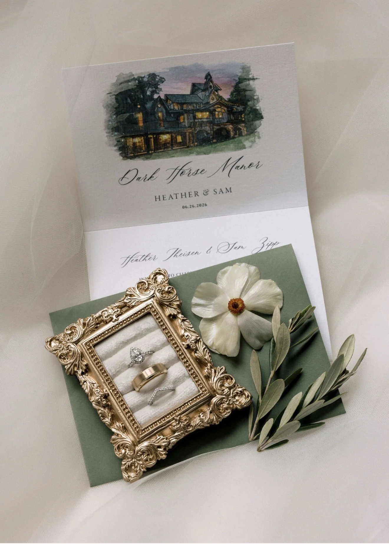 A flat lay of a wedding invitation for a wedding in Franklin, Tennessee. Atop the invitation sits a box of rings, a white flower, and some greenery.