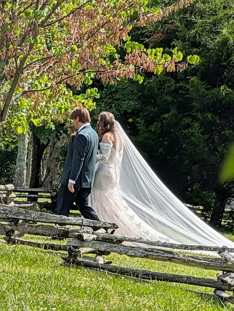 A bride and groom walking outdoors under trees with pink and green leaves, on grassy terrain with a rustic wooden fence.