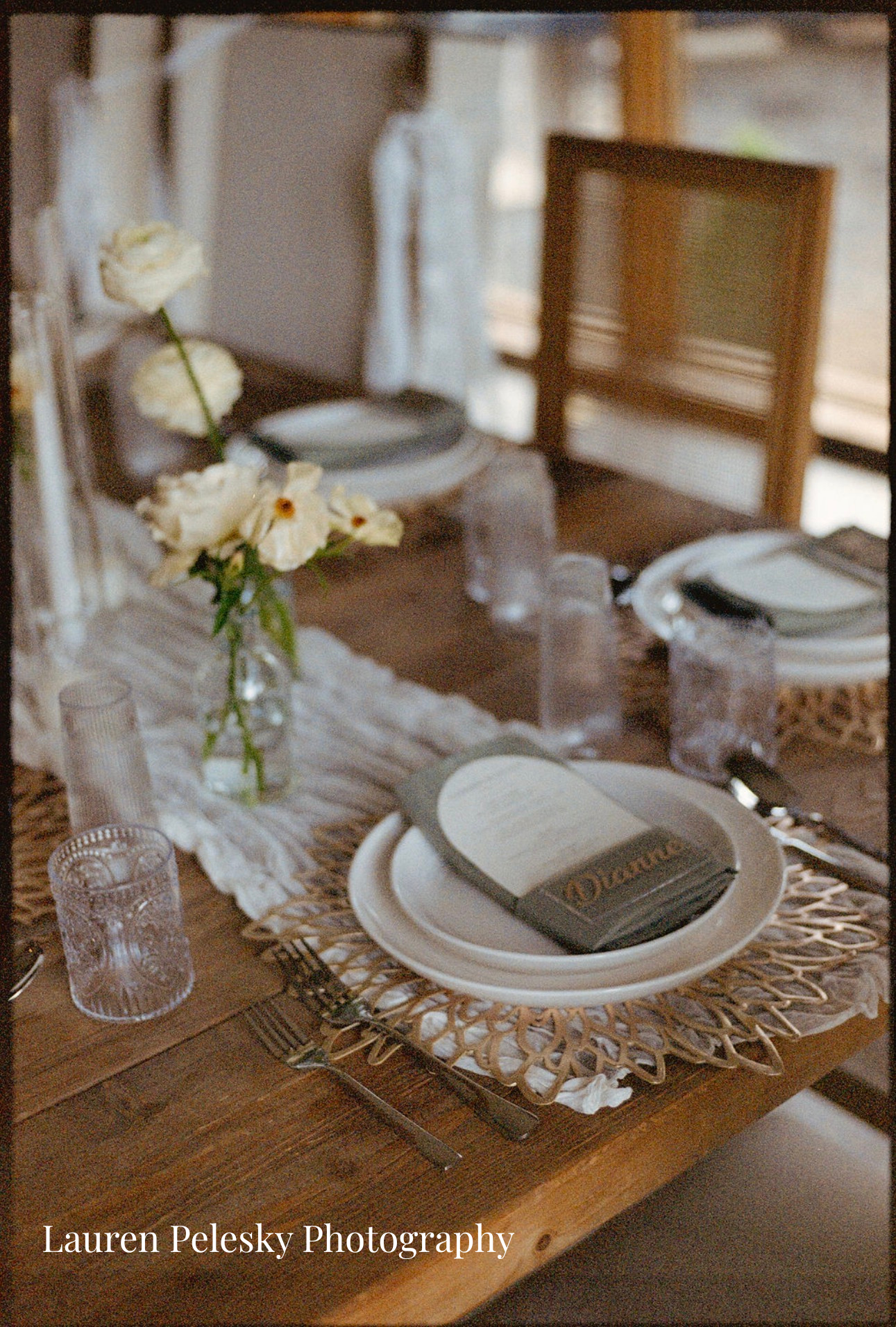 A rustic wooden dining table set for a meal with a woven placemat, white plate, gray napkin, black cutlery, and a hardcover book on top. There are clear textured drinking glasses, a small glass vase with white flowers, and additional glasses on the t