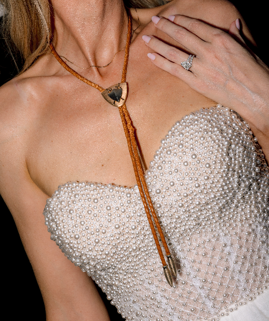 A bride wearing a pearl studded dress shows off her bolo-tie at a farewell party for a wedding in Franklin, Tennessee.