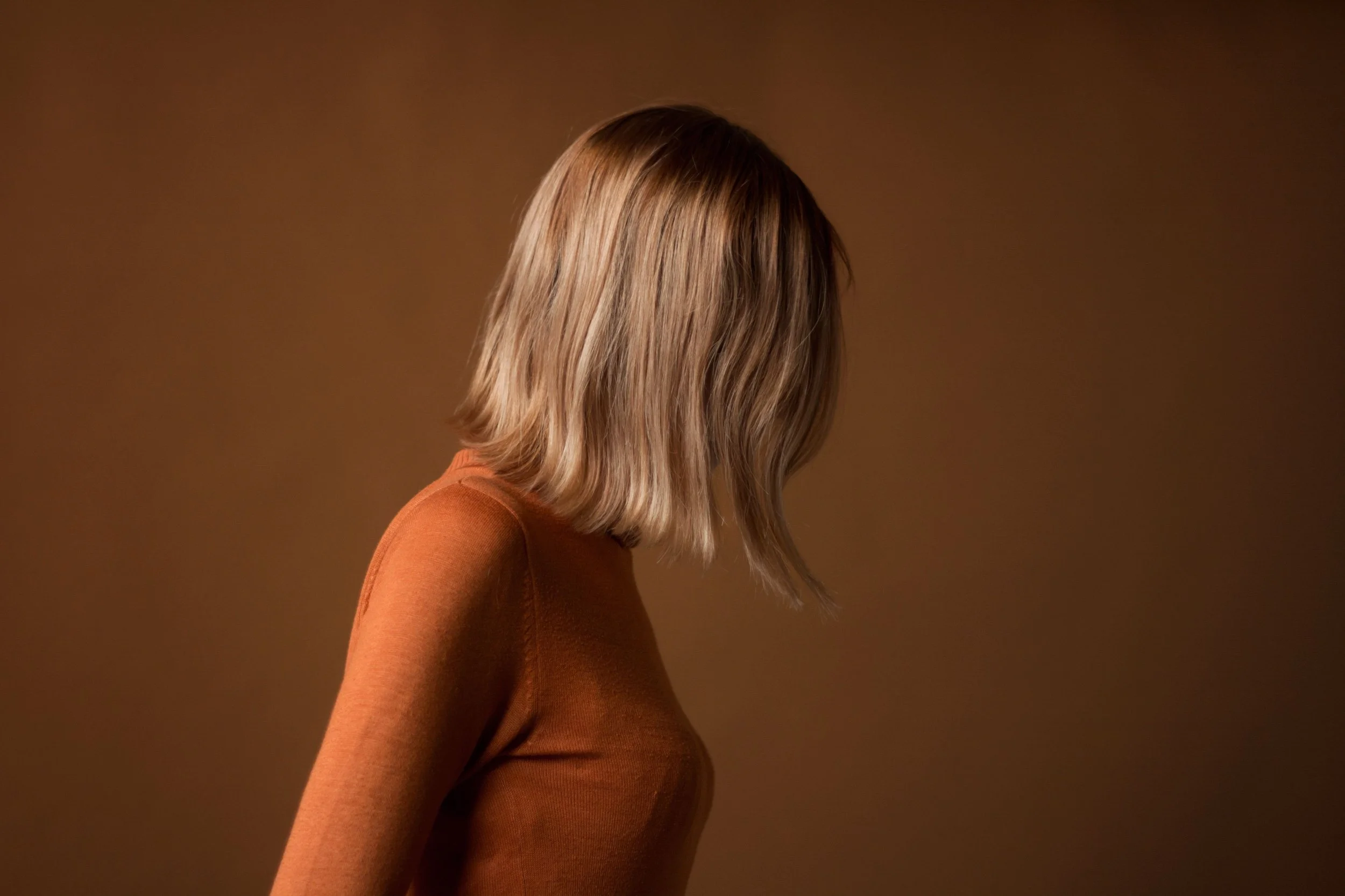 Portret van een vrouw met schouderlange, golvende blondine haren, gekleed in een oranje trui, zichtbaar vanaf de zijkant tegen een effen, bruinachtige achtergrond.