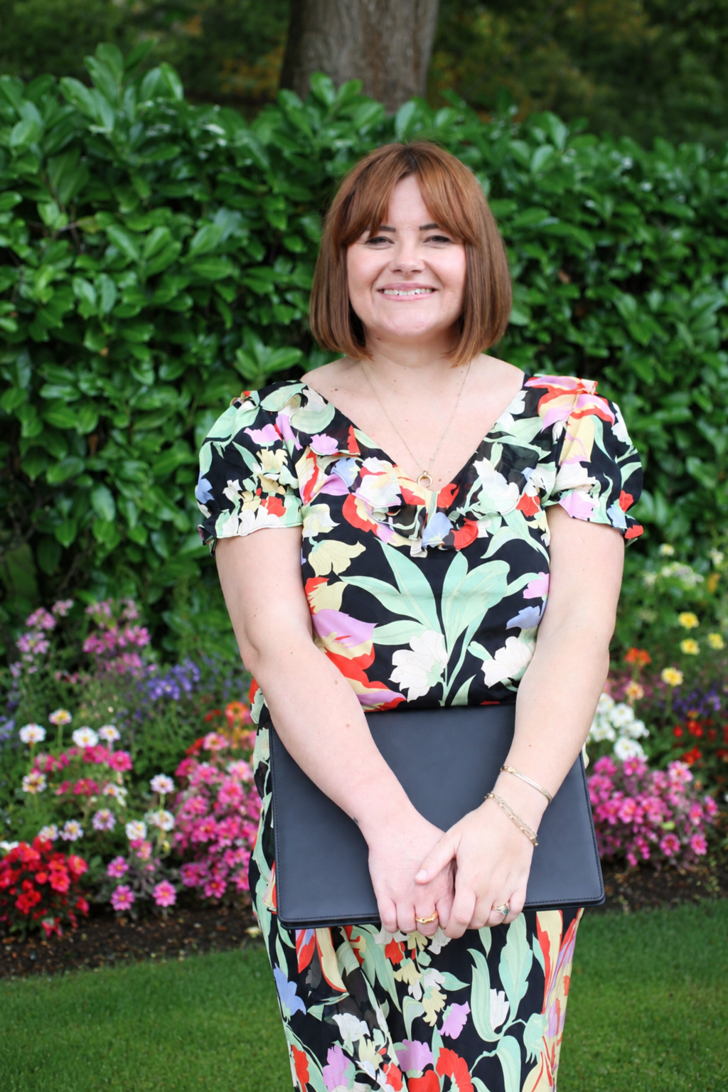 A woman with shoulder-length red hair, smiling, standing outdoors in front of green bushes and flowering plants, holding a black portfolio.