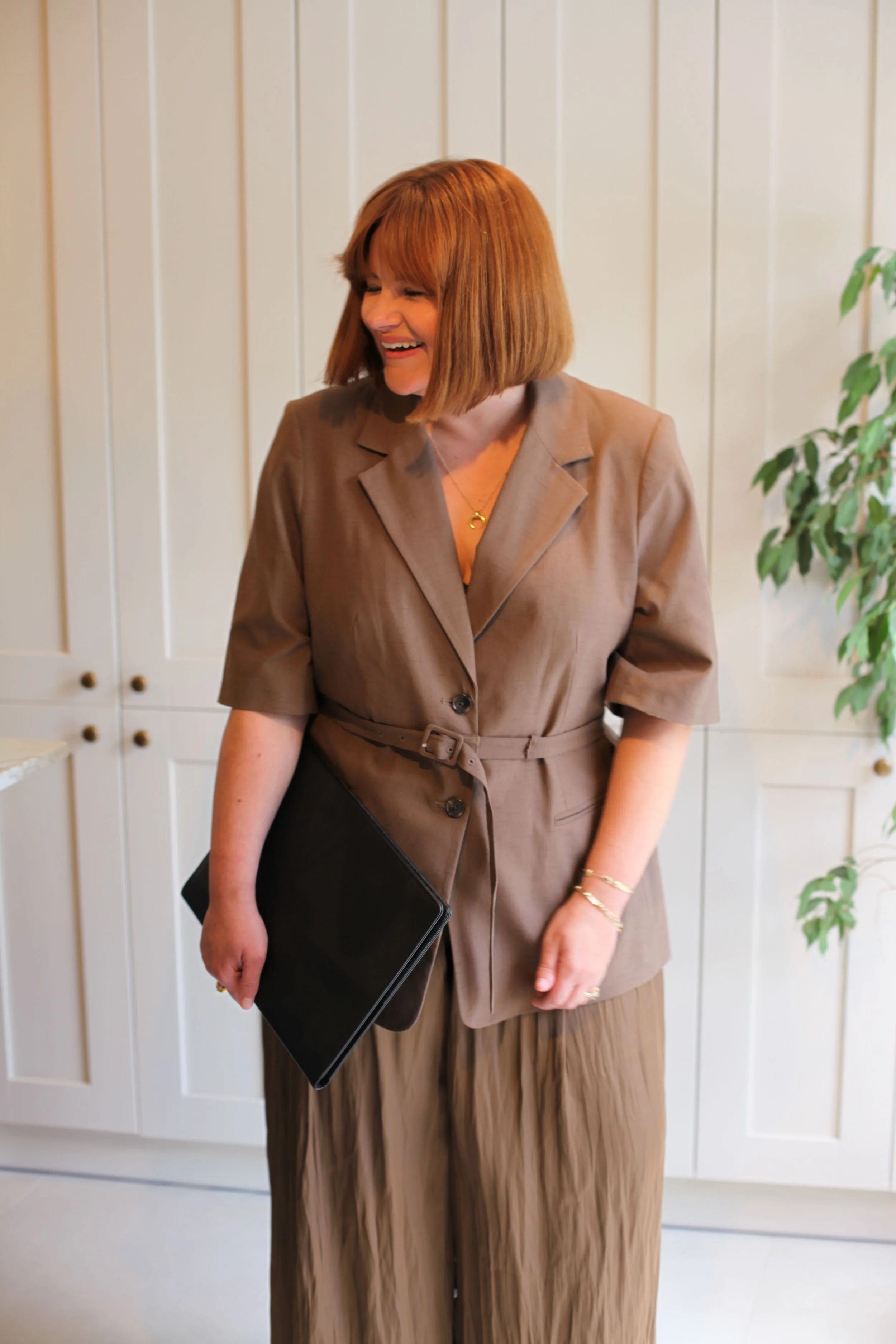 A woman with shoulder-length red hair wearing a brown blazer and matching pleated skirt holding a black folder, standing in front of white cabinets with gold knobs, and a green plant on the right.