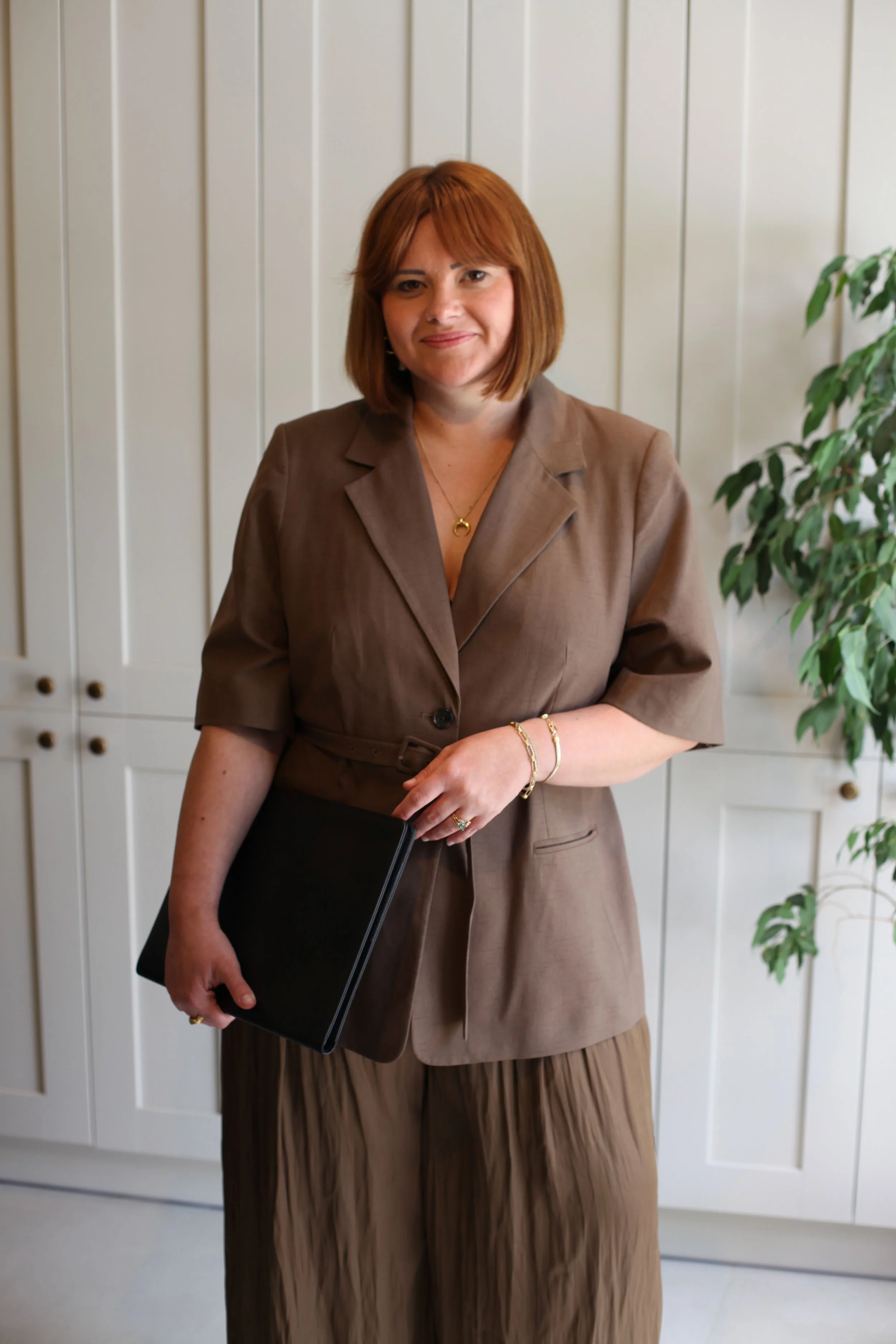 A woman with shoulder-length reddish-brown hair, wearing a brown blazer and matching skirt, holding a black folder, and standing in front of white paneled cabinets and a potted green plant.
