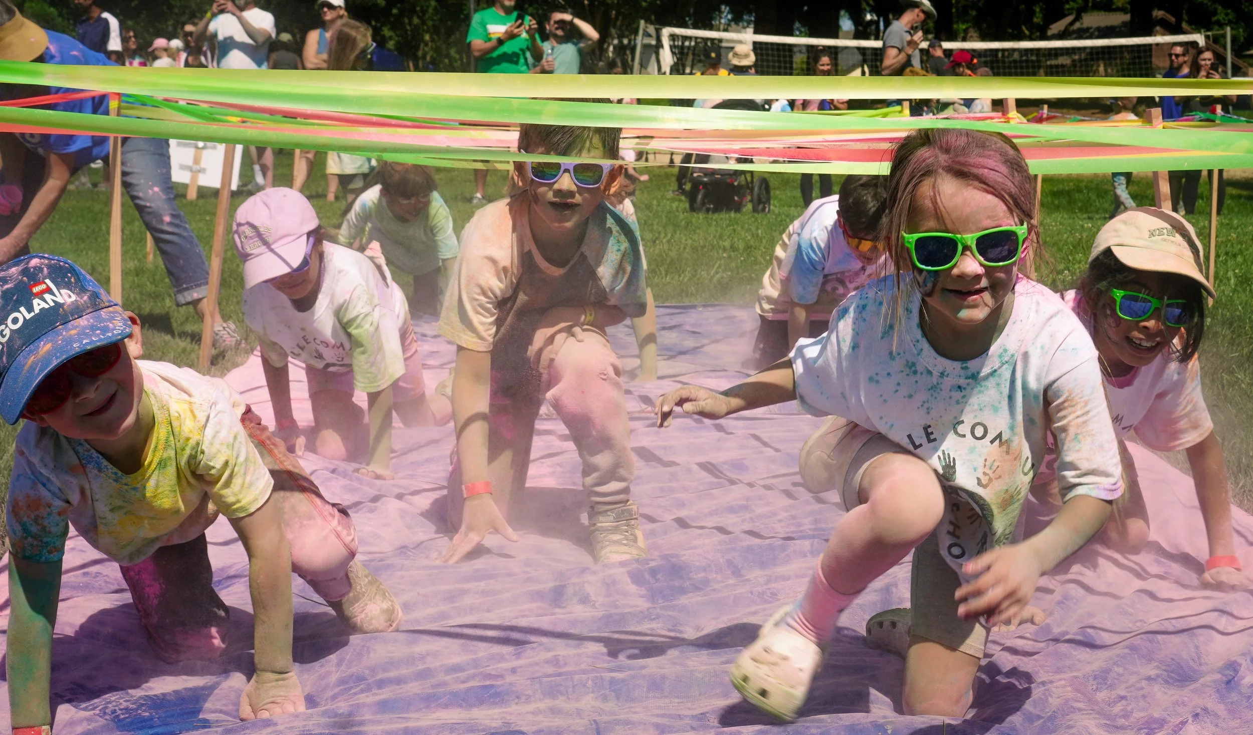 Children participating in a color run event, crawling under colorful streamers on a pink tarp, with spectators in the background.