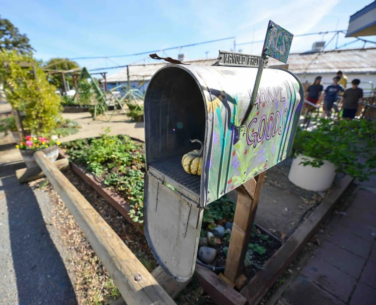 Decorative mailbox with signs reading 'GARDEN GOODS', painted with colorful and floral designs, located in a community garden with plants and people in the background.