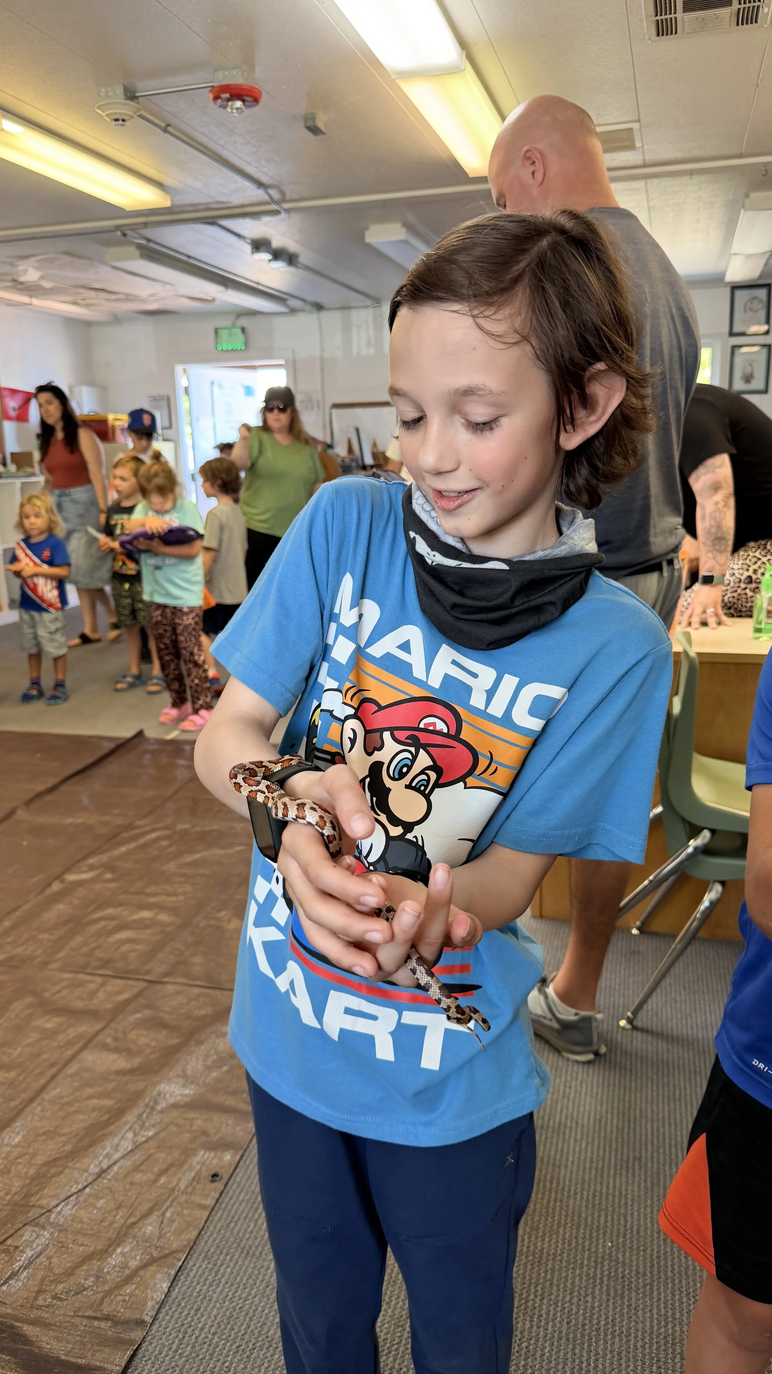 A young boy in a blue t-shirt with a Mario design, holding a small snake. He is smiling and looking down at the snake. In the background, there are several other children and adults in a room with bright lighting and framed pictures on the wall.