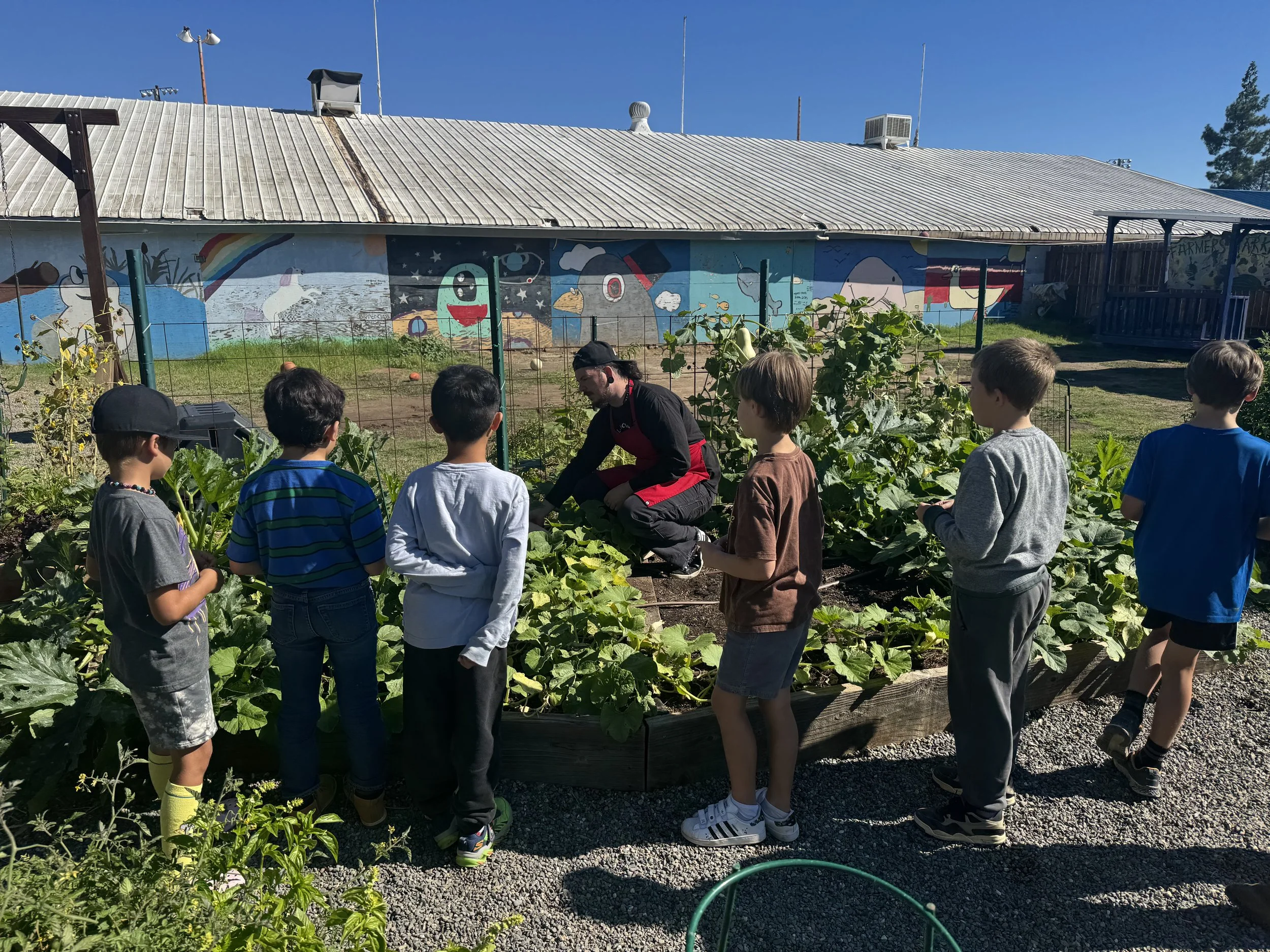 A group of children gathered around a woman in a garden, with a building featuring colorful murals on the wall behind them. The mural depicts animals, a rainbow, and clouds. The setting appears to be a sunny day.