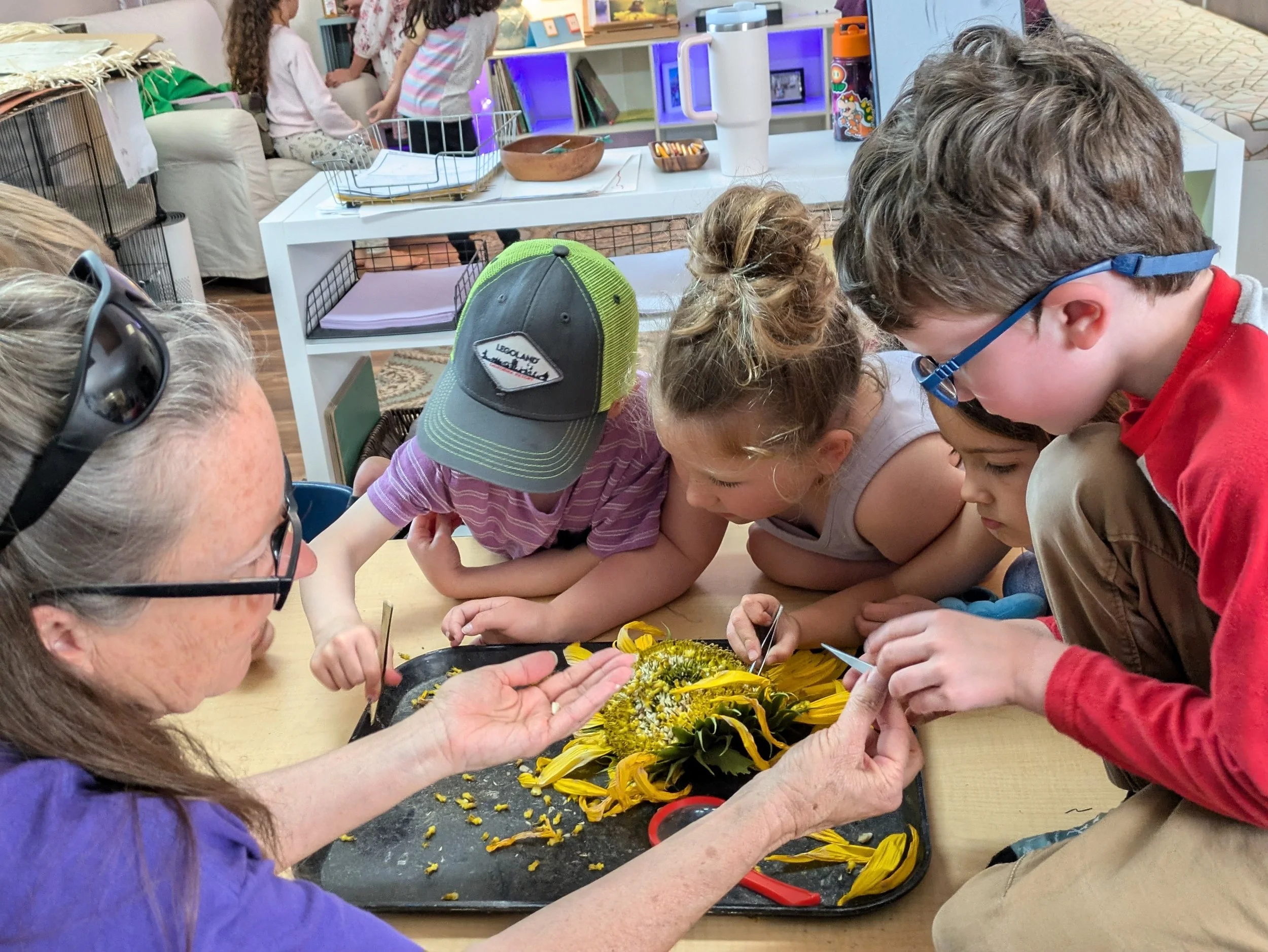 A group of children and an adult gathered around a table, working on a craft project involving yellow flowers and leaves, with various supplies and decorations in the background.