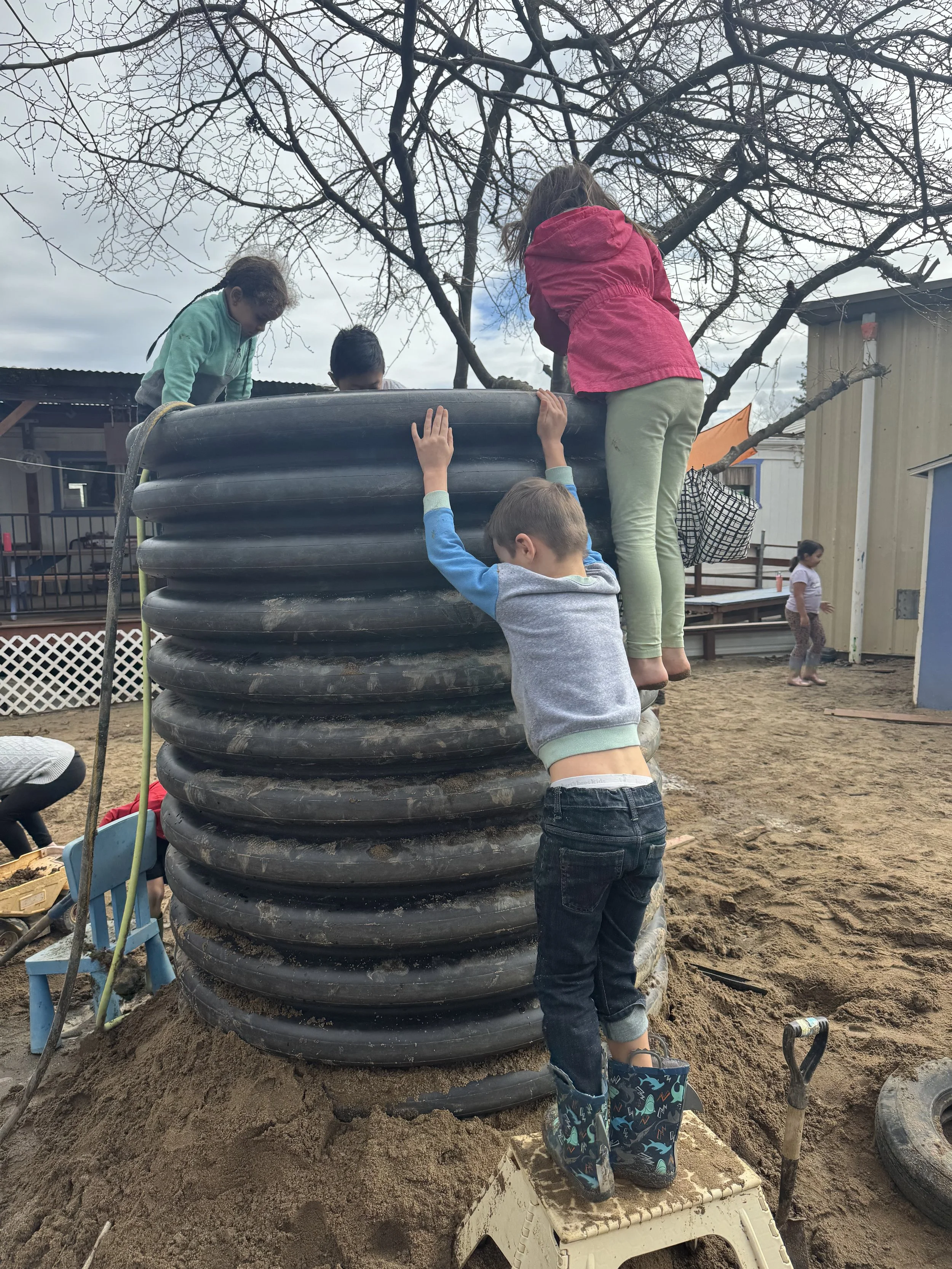 Children playing on a construction site with a large black tire wall climbing structure, some children are climbing while one is standing on a small platform at the base.