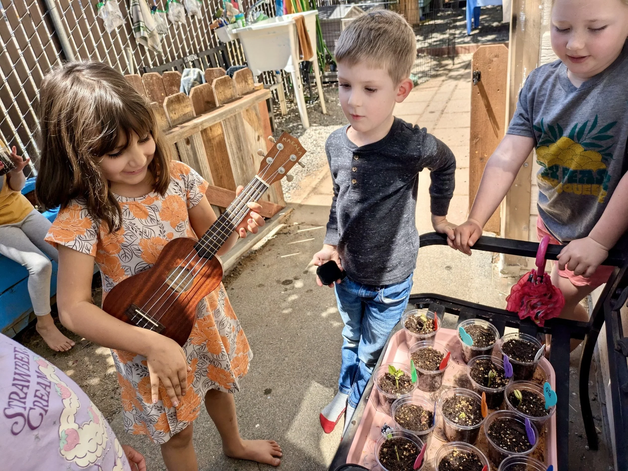Three children are outside near a table with small plant pots. One girl with brown hair and a floral dress is holding a ukulele. A boy with short light brown hair and a dark gray shirt is looking at the table. Another girl with blonde hair, a gray shirt, and pink shoes is also looking at the plant pots. The table has multiple small pots with soil and plants, some labeled with colorful tags.