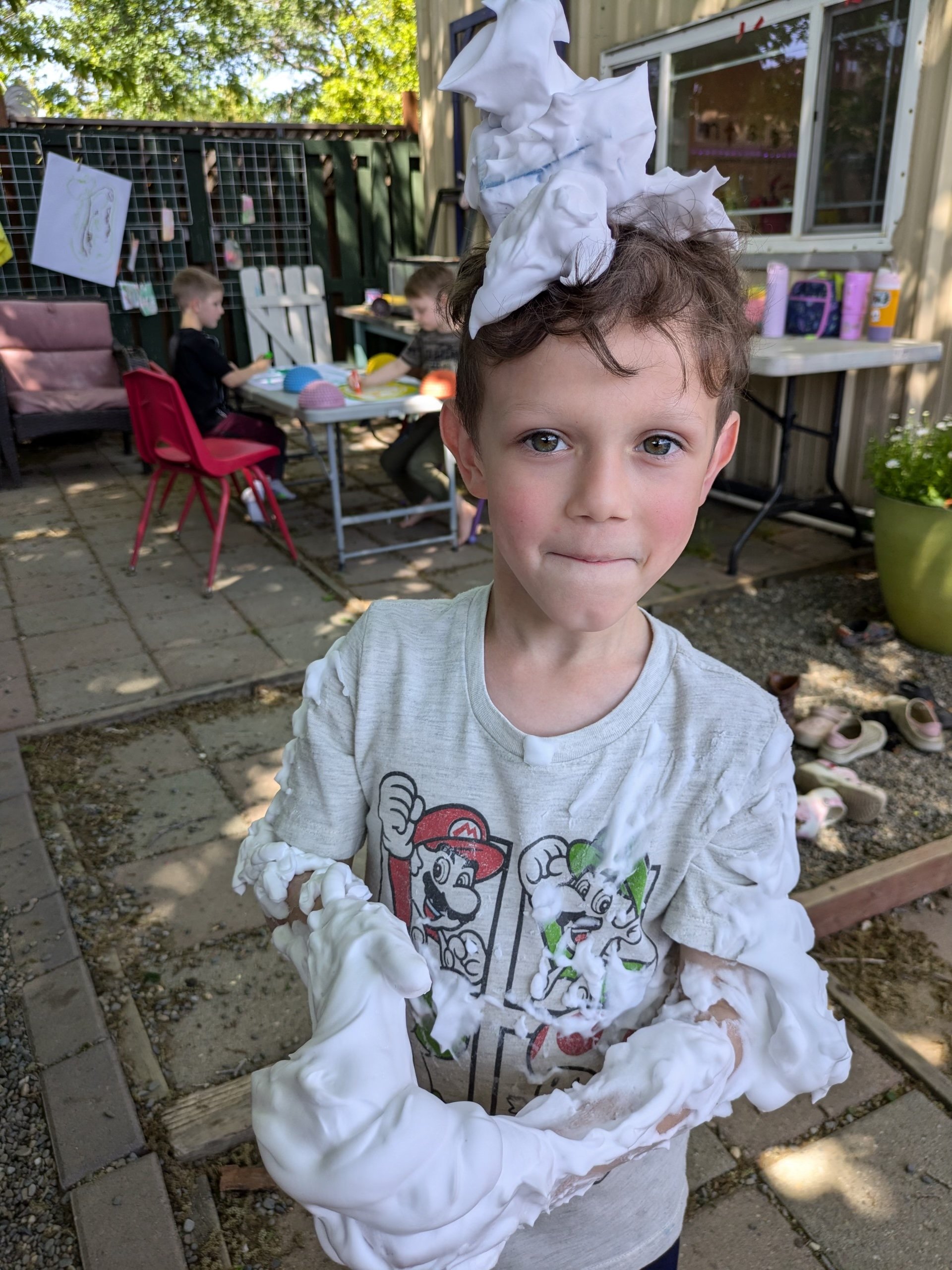 Young boy with brown curly hair and light skin, covered in white foam, wearing a white long-sleeve shirt with Mario graphics, standing outdoors on a sunny day.