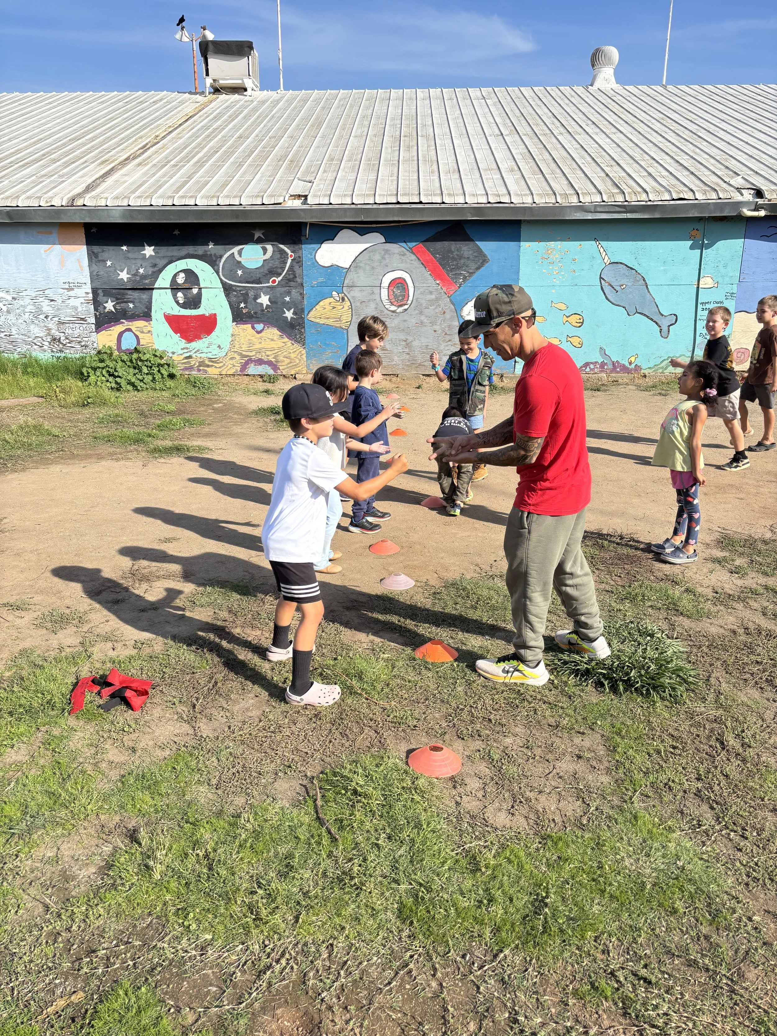 Children and an adult standing outdoors near a colorful mural, engaging in a game involving orange and pink cones, on a sunny day with a dirt and grass ground.