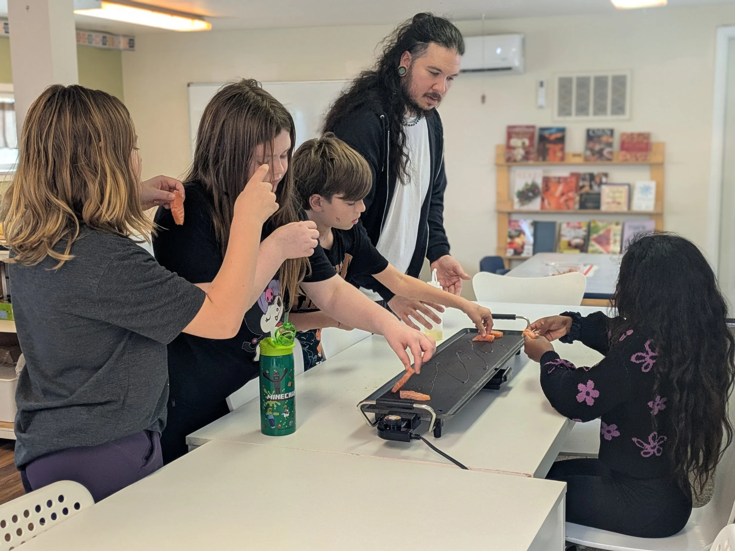 A group of children and an adult in a classroom are gathered around a table with a baking tray, making snacks. One child is holding a piece of salmon, and the adult is assisting. Other children are watching and participating. Books and educational materials are visible in the background.