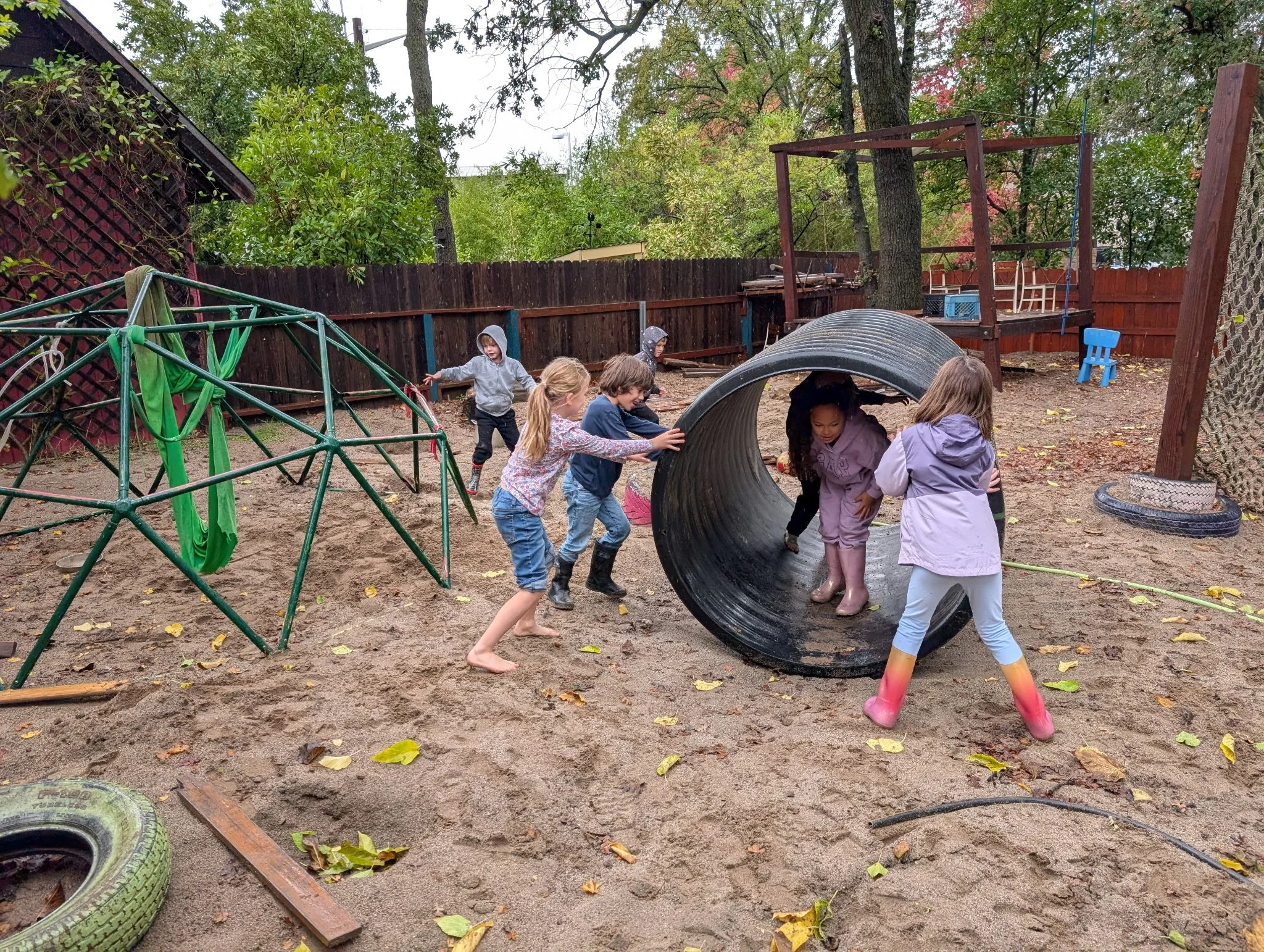Children playing in a backyard playground with a large black tube tunnel, a climbing structure with green ropes, and a wooden platform with chairs, surrounded by trees and a wooden fence, on an autumn day.