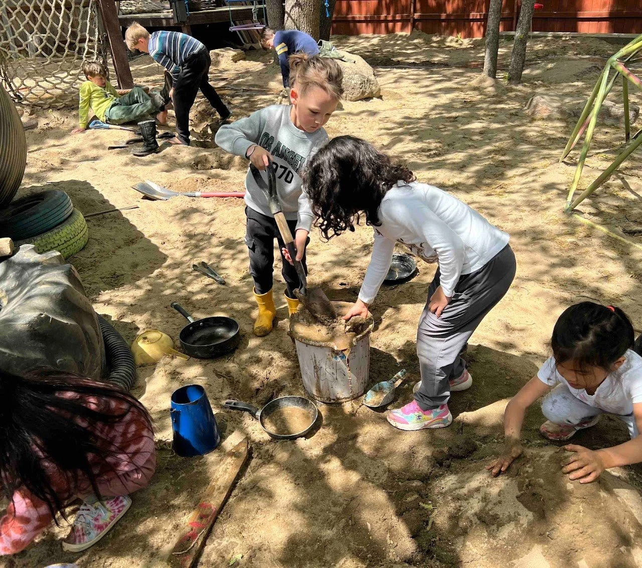 Children playing and digging in a sandbox with shovels and toys, some wearing rubber boots, surrounded by trees and sand, with a wooden fence in the background.
