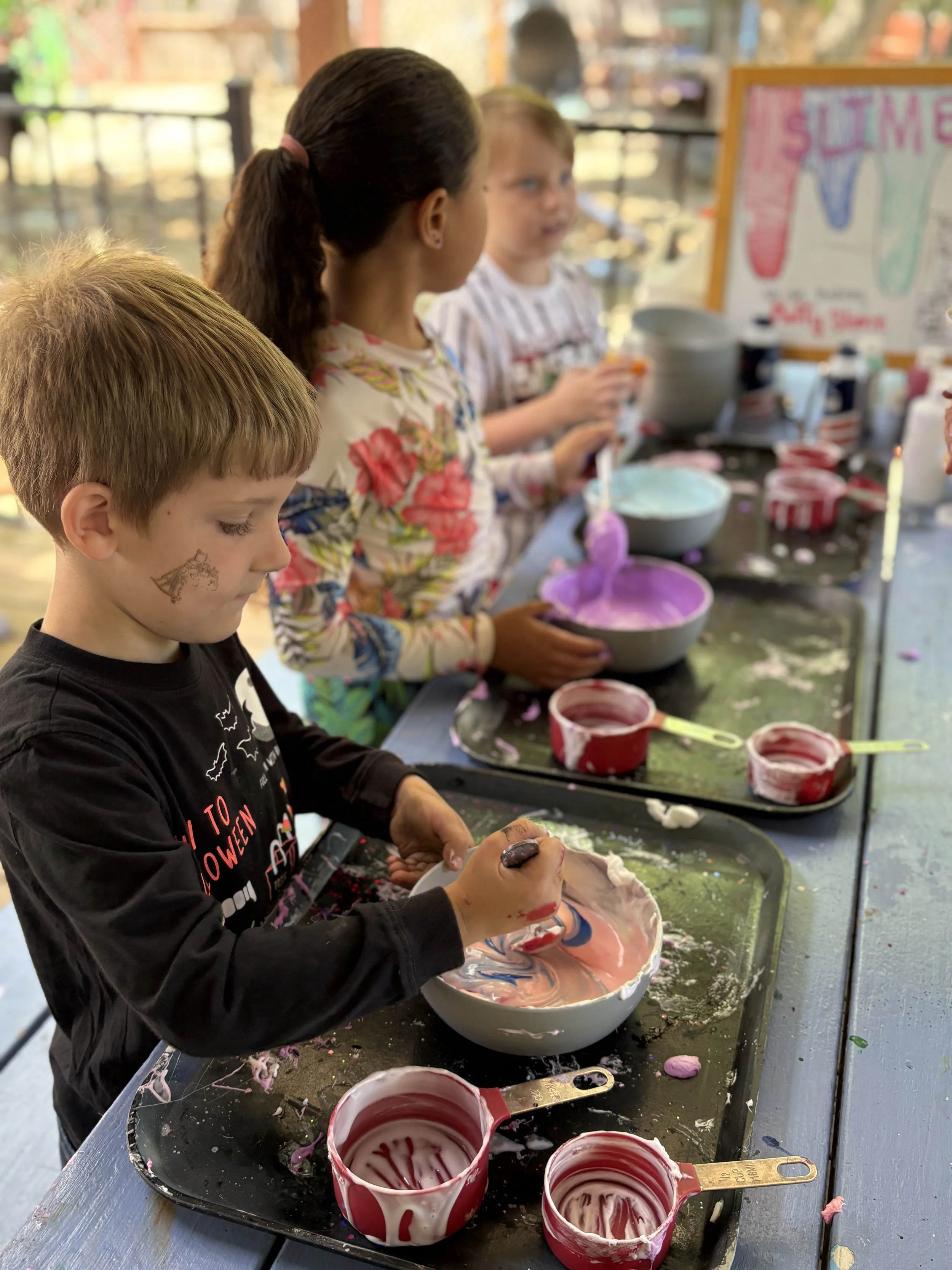 Children participating in a tie-dye activity at an outdoor table, with bowls of colorful dye and a sign in the background.