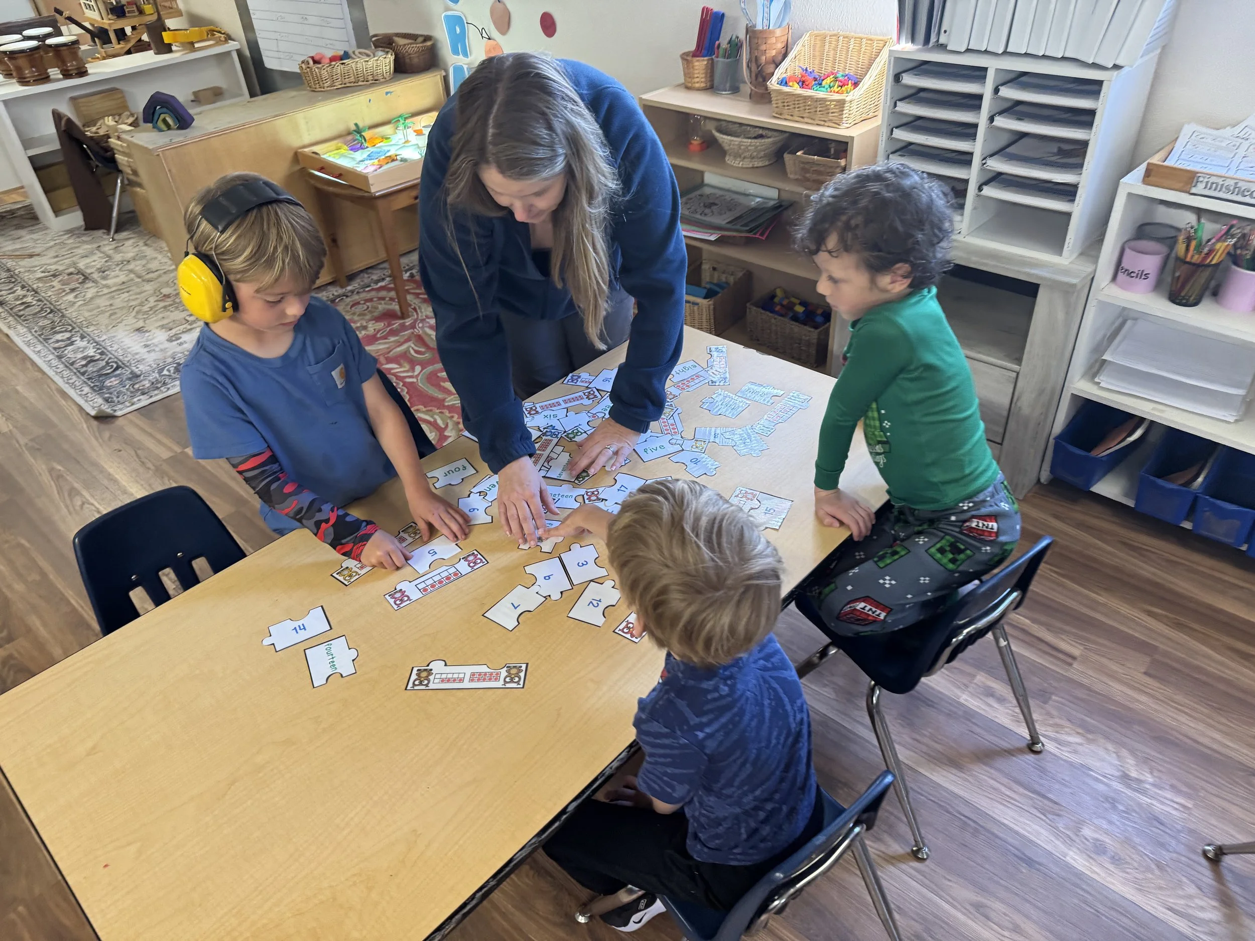 A teacher and four young children are gathered around a table playing an educational puzzle game with number pieces and images in a classroom.