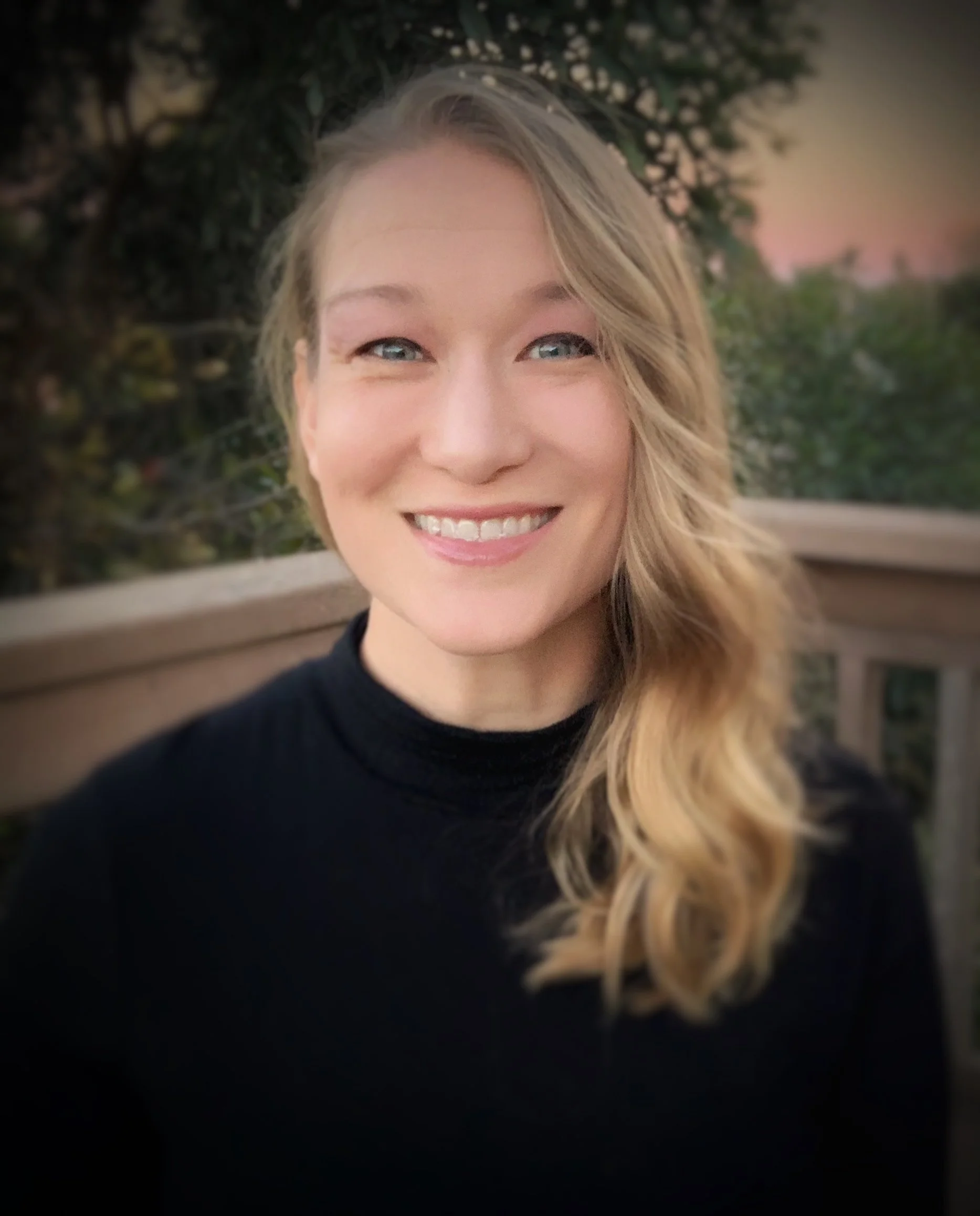A photo of Dr. Buttery - a smiling blonde woman with blue eyes outdoors at sunset, with trees and sky in the background.