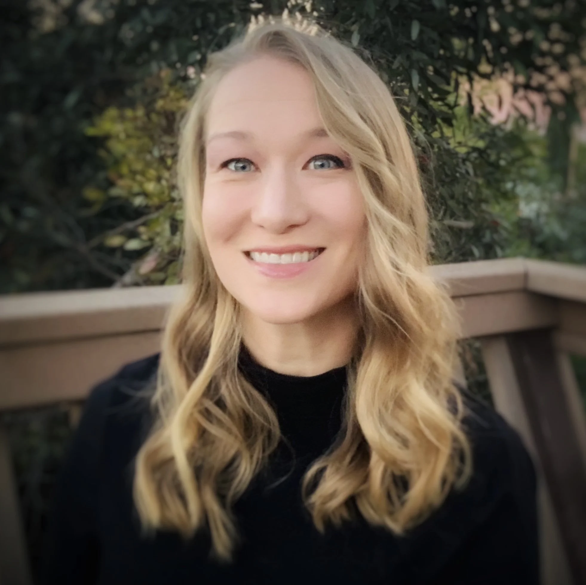 Close-up of a young woman with blonde wavy hair, smiling, with blurred greenery in the background.