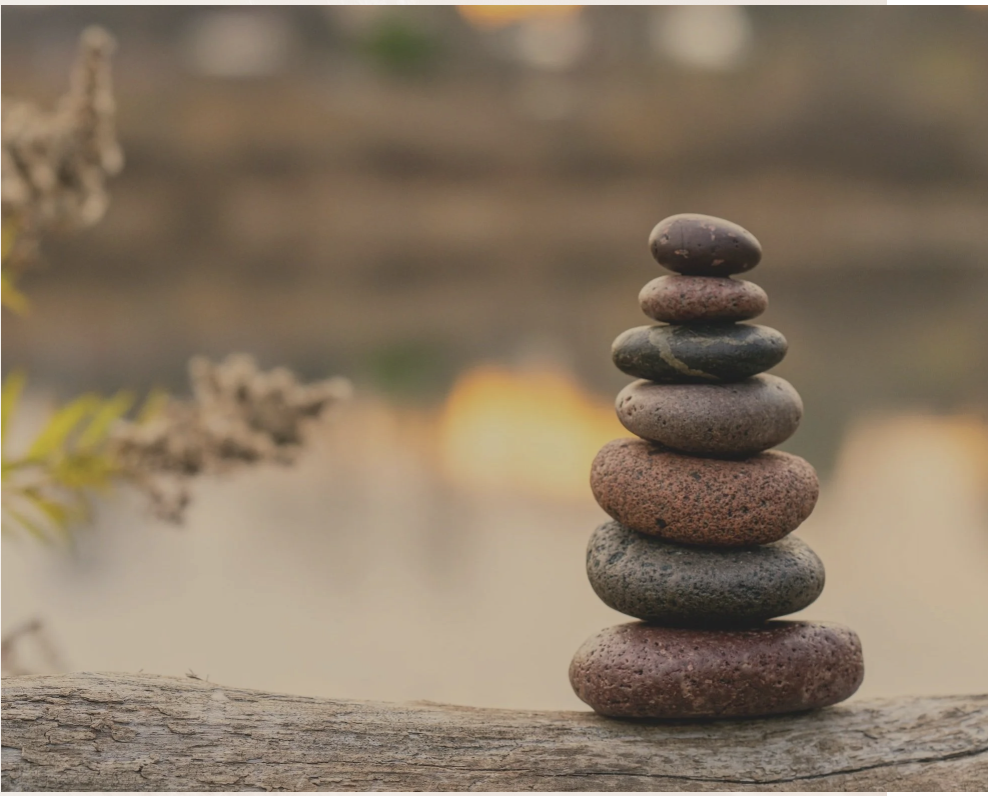 Stack of eight smooth, rounded stones of various colors balanced on a piece of driftwood near water with blurred background.