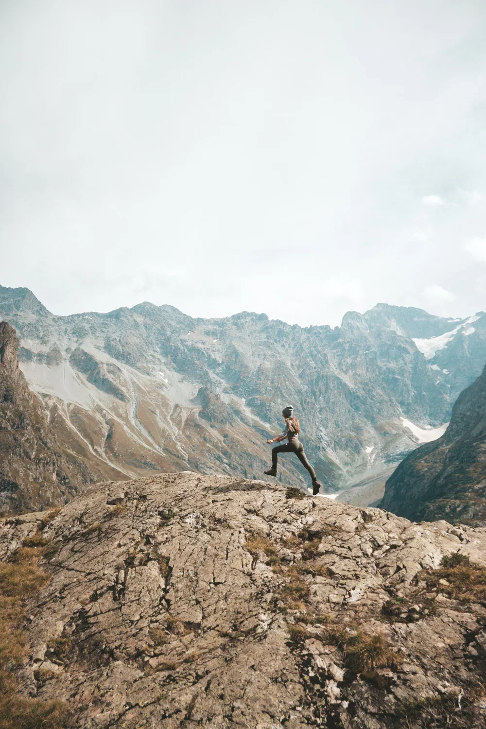 A person wearing a helmet and outdoor clothing running on a rocky mountain terrain with rugged mountains and partly cloudy sky in the background.