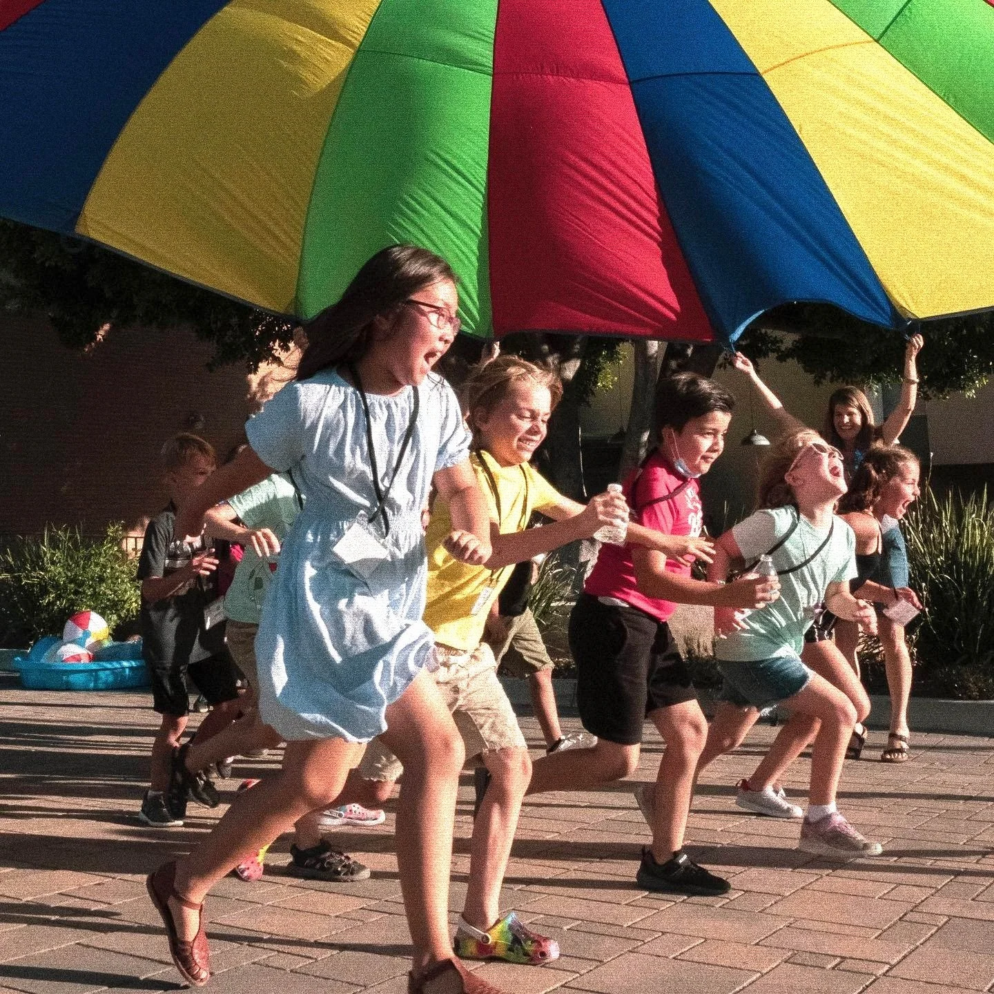 Group of children running outdoors under a large colorful umbrella on a sunny day.