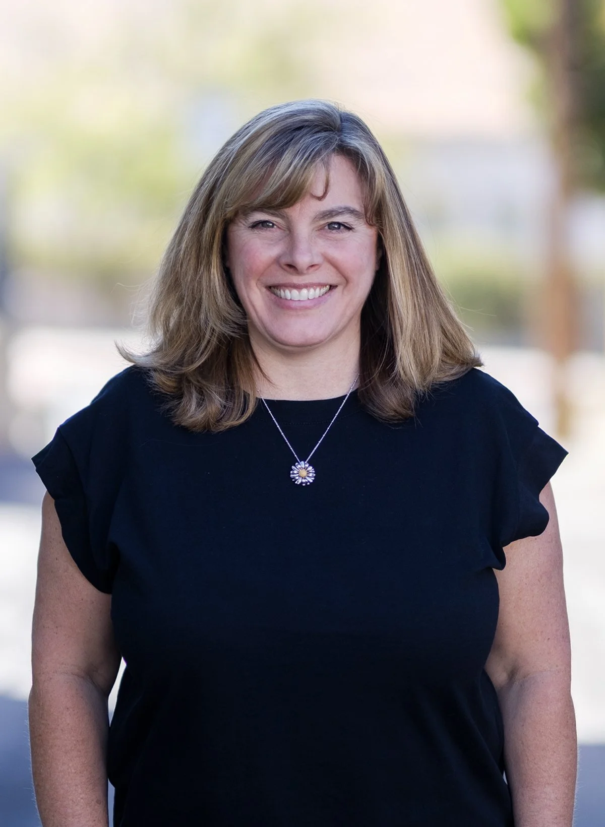 A woman with shoulder length light brown hair smiling outdoors, wearing a black top and a flower-shaped pendant necklace.