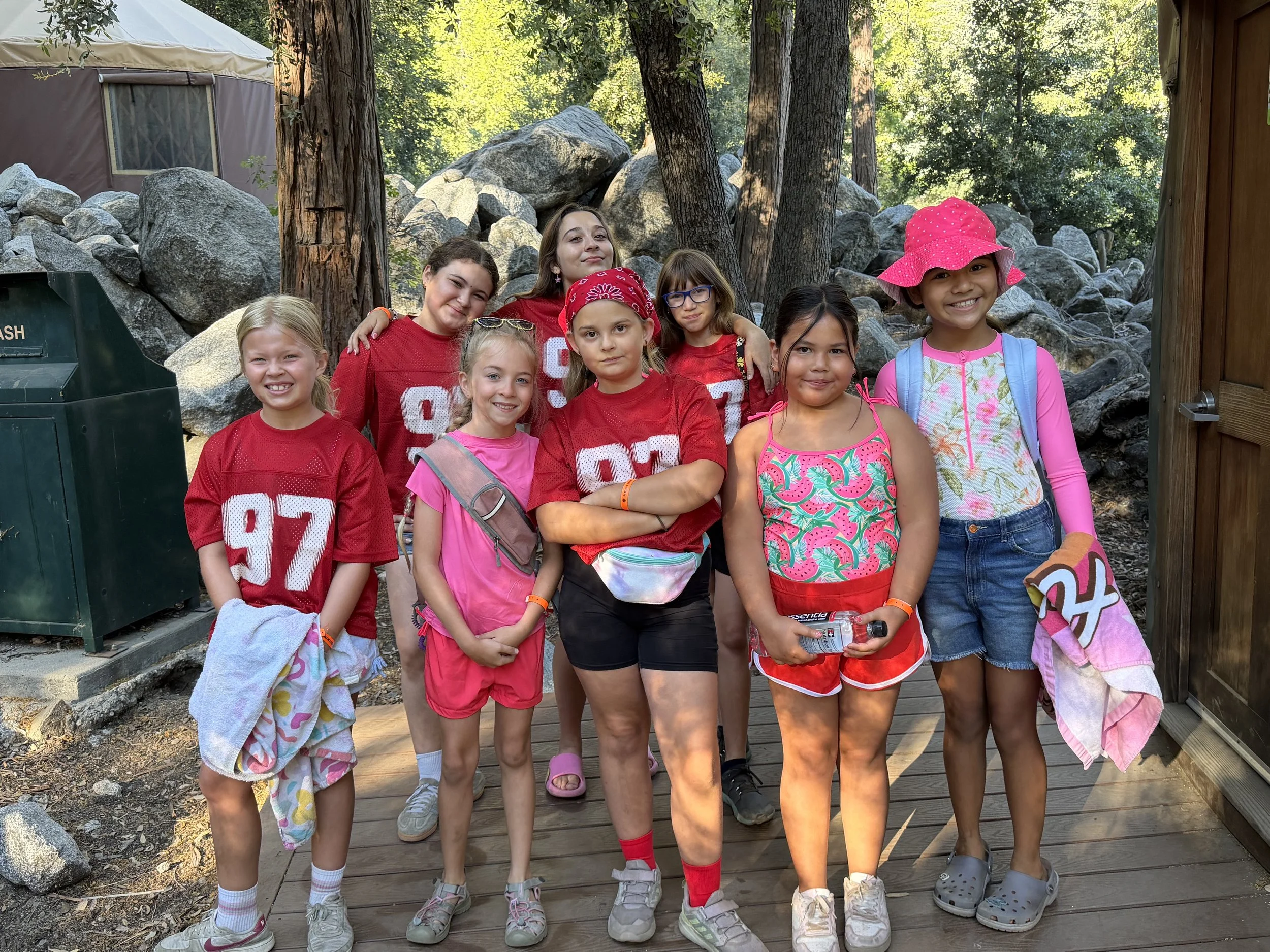 Group of nine young girls with diverse appearances standing outdoors on a wooden pathway, smiling, wearing colorful casual summer clothes, with trees and rocks in the background.