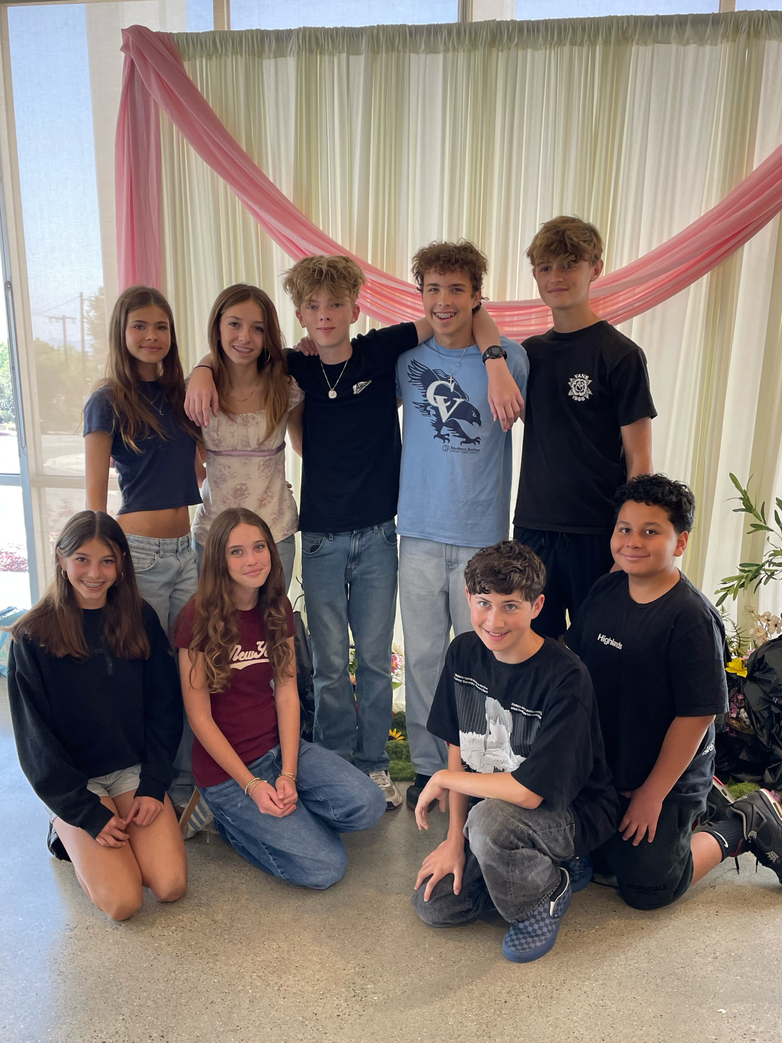 Group of teenagers posing for a photo indoors, with cream and pink drapery in the background. Some are kneeling, and some are standing, smiling at the camera.