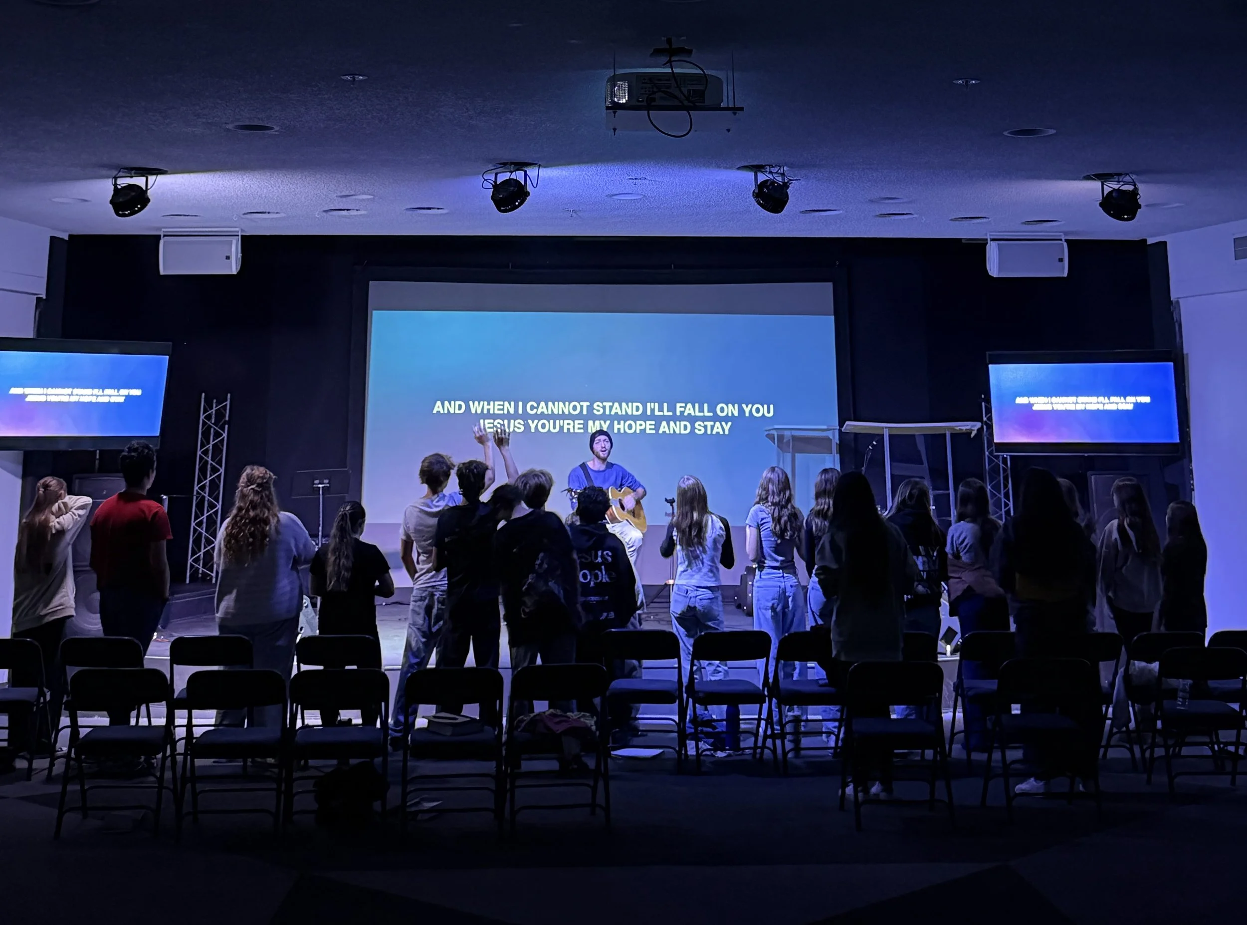 A group of people attending a church service or worship session in a dark room with a stage, a large screen displaying song lyrics, and a musician playing guitar in front of the congregation.